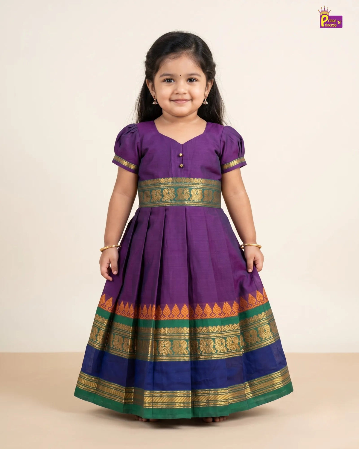 A young girl modeling a purple traditional Indian dress with gold and green borders, standing against a plain beige background.