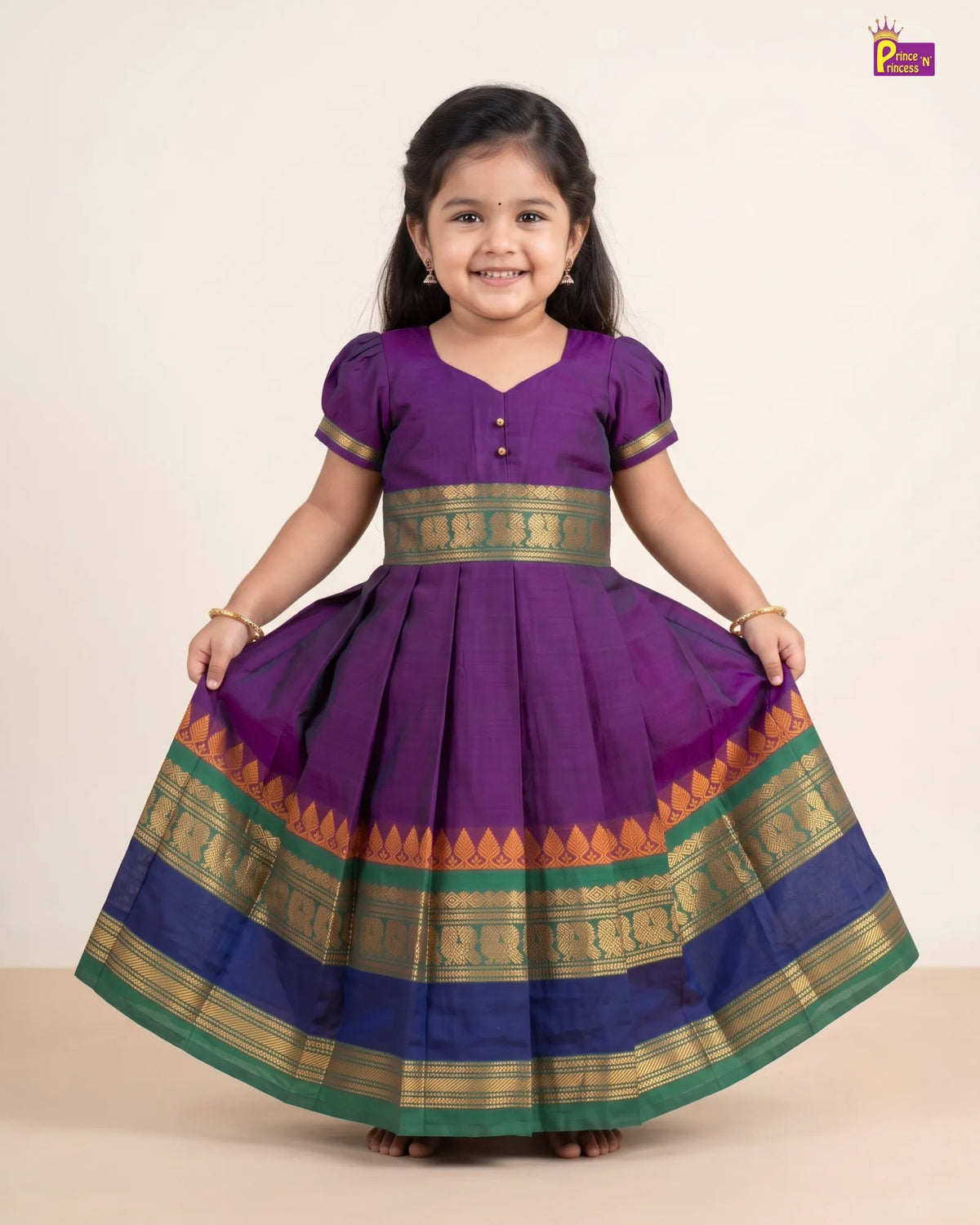A young girl smiling and holding out a purple traditional dress with gold, green, and blue borders, photographed against a plain beige studio background.