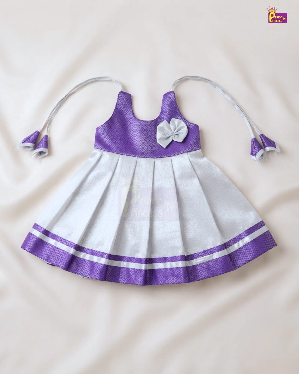 A baby girl's sleeveless dress with a purple textured bodice, white pleated skirt, a white bow on the chest, and purple and white striped trim at the hem. The dress has thin shoulder straps that extend into bow-tie hair clips.