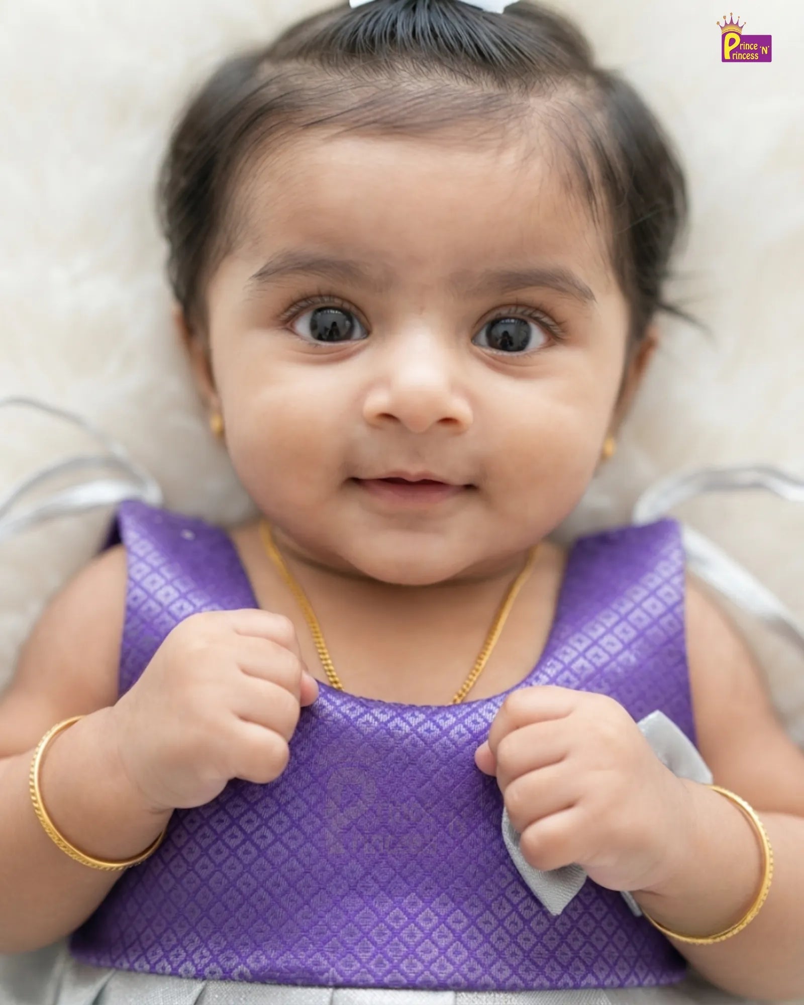 A close-up of a baby wearing a purple patterned sleeveless top, gold jewelry, and a small gray bow, lying on a soft light background.