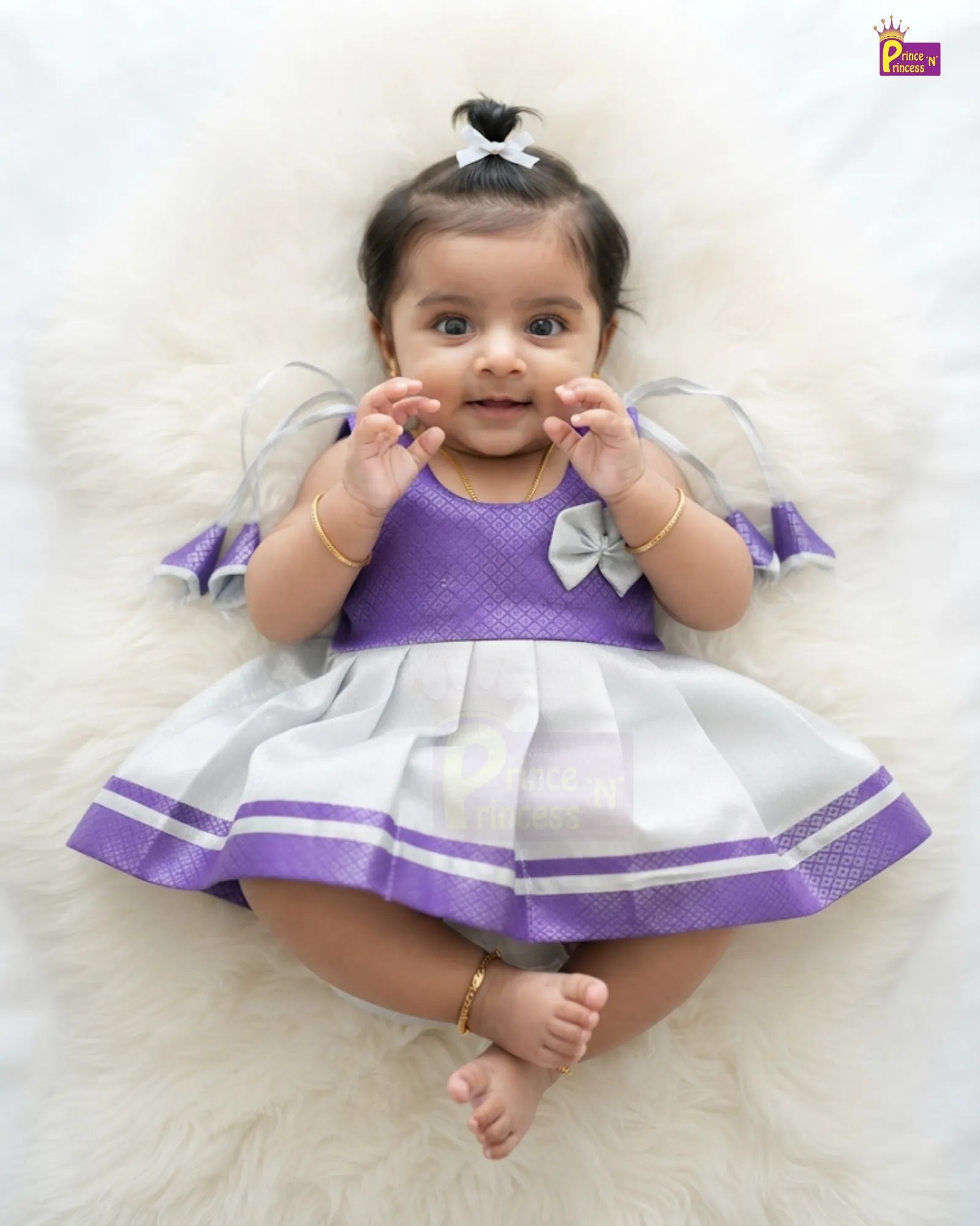 A baby girl wearing a purple and white dress, lying on a white faux fur rug. The dress has a purple textured bodice with a white bow, and a white pleated skirt with purple stripes and ruffled sleeves.