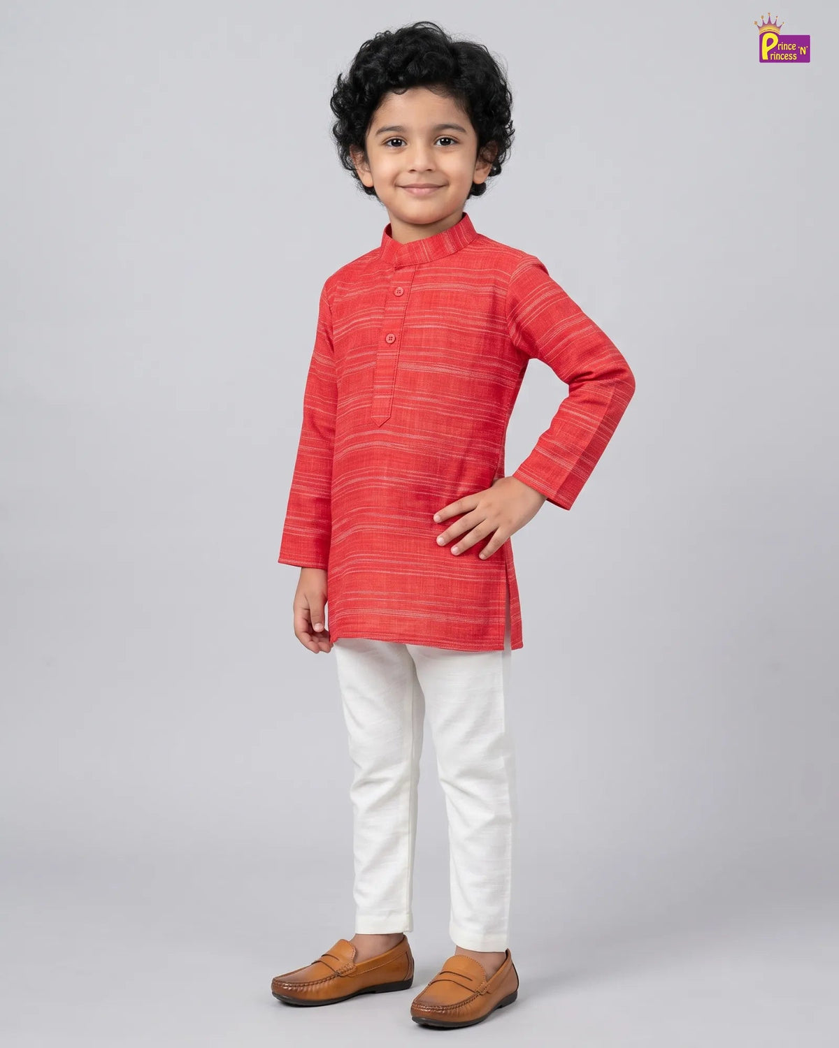 A young boy modeling a red and orange striped kurta with white trousers and brown shoes, standing against a light gray studio background.
