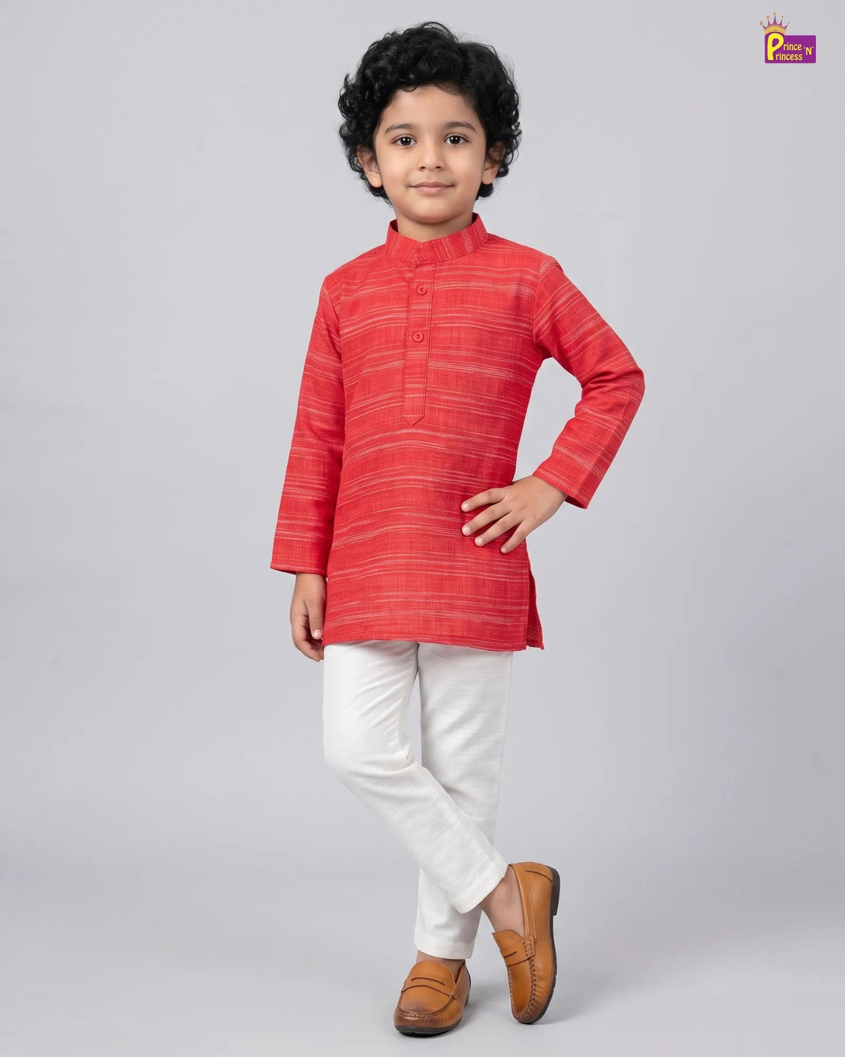 A young boy modeling a red and orange striped kurta with white trousers and brown shoes, standing against a light gray studio background.