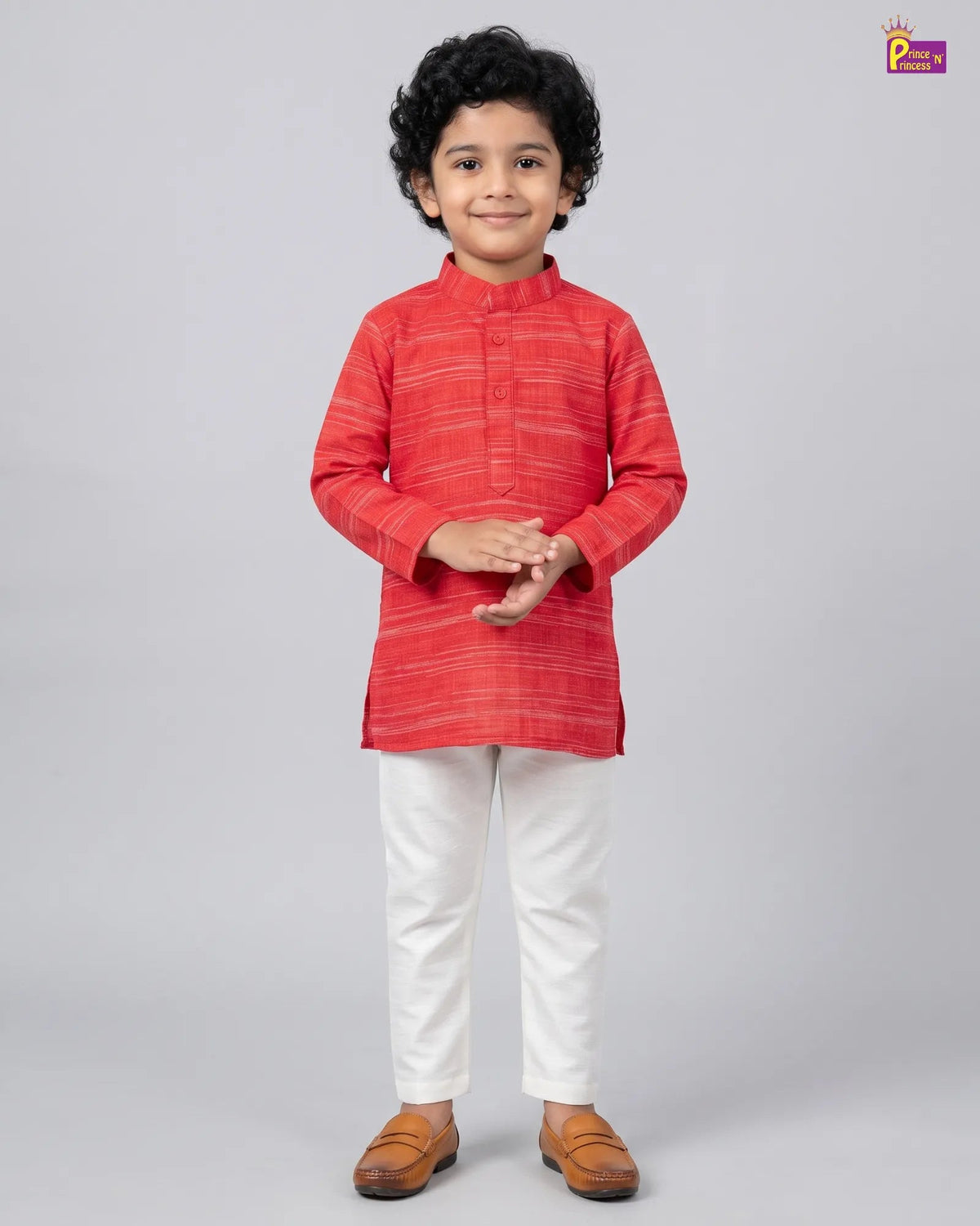 A young boy modeling a red and orange striped kurta with white trousers and brown loafers, standing against a solid gray studio background.
