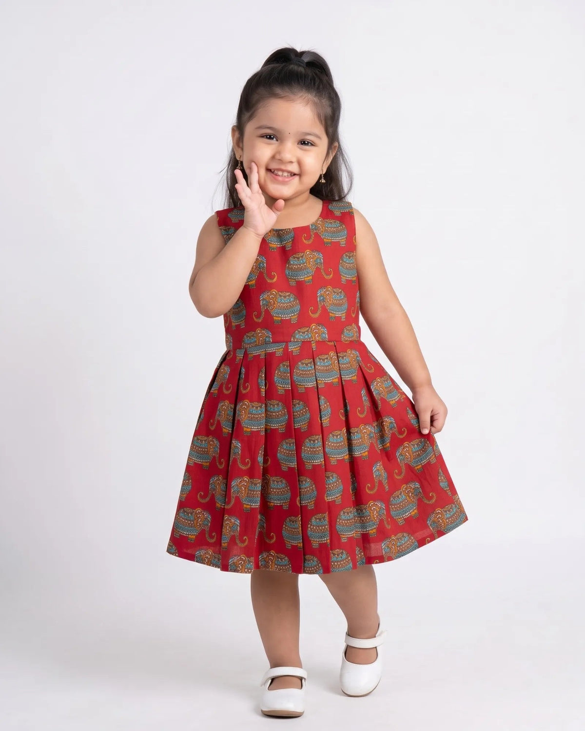 A young girl with her hair in a high ponytail is wearing a sleeveless red dress with a green and gold elephant print, posing against a plain white studio background.