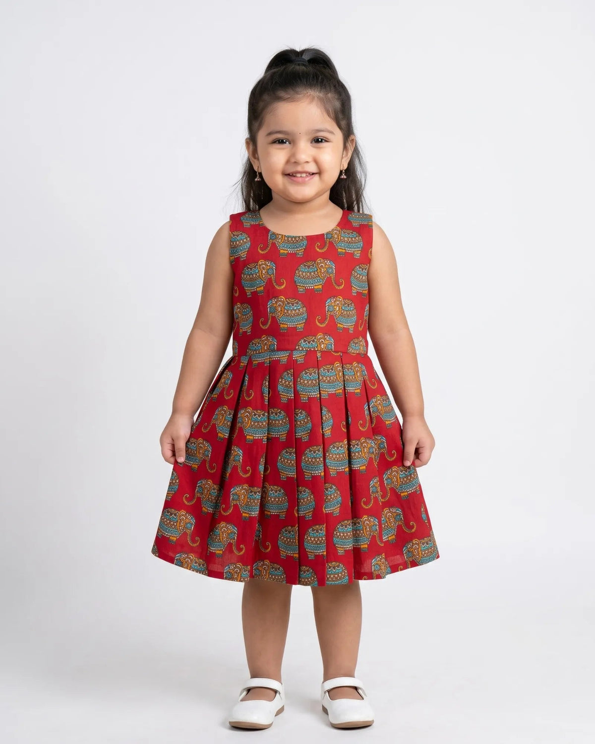 A young girl with her hair in a high ponytail stands against a light gray studio background wearing a sleeveless red dress with a repeating elephant print in blue, green, and yellow, paired with white Mary Jane shoes.