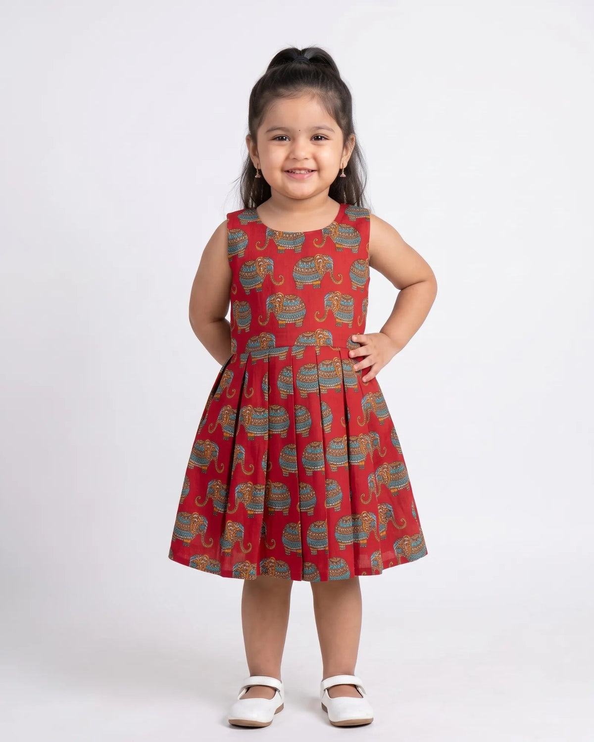 A young girl with her hair in a high ponytail stands against a light gray studio background wearing a sleeveless red dress with a repeating elephant print in blue, green, and gold, paired with white Mary Jane shoes.