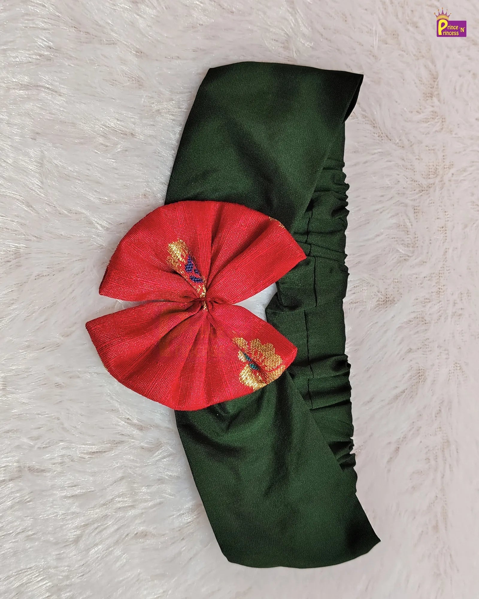 A dark green fabric headband with a large red bow featuring gold and purple embroidered details, laid flat on a white faux fur background.