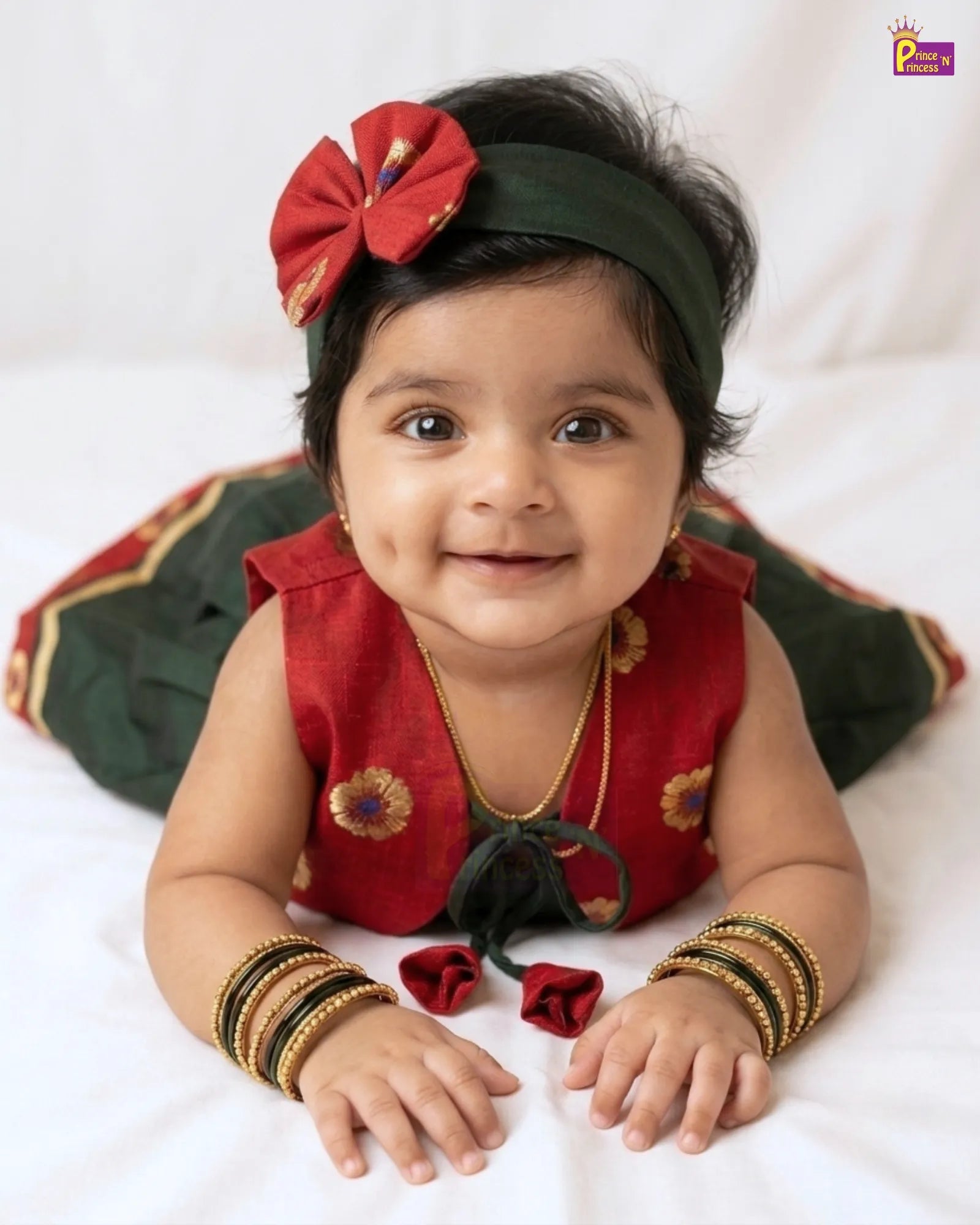 A baby girl wearing a traditional Indian outfit, featuring a red sleeveless top with gold floral embroidery, a dark green headband with a red bow, and gold bangles, lying on a white fabric background.