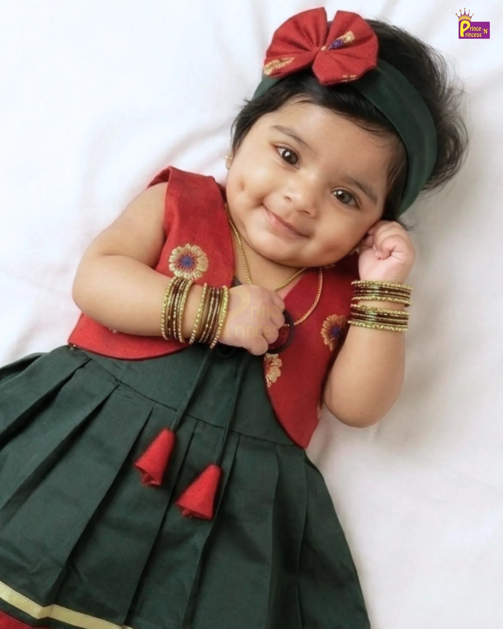 A baby girl lying on a white sheet, wearing a traditional Indian outfit with a dark green pleated skirt, a red sleeveless vest, a matching red bow headband, and gold bangles.