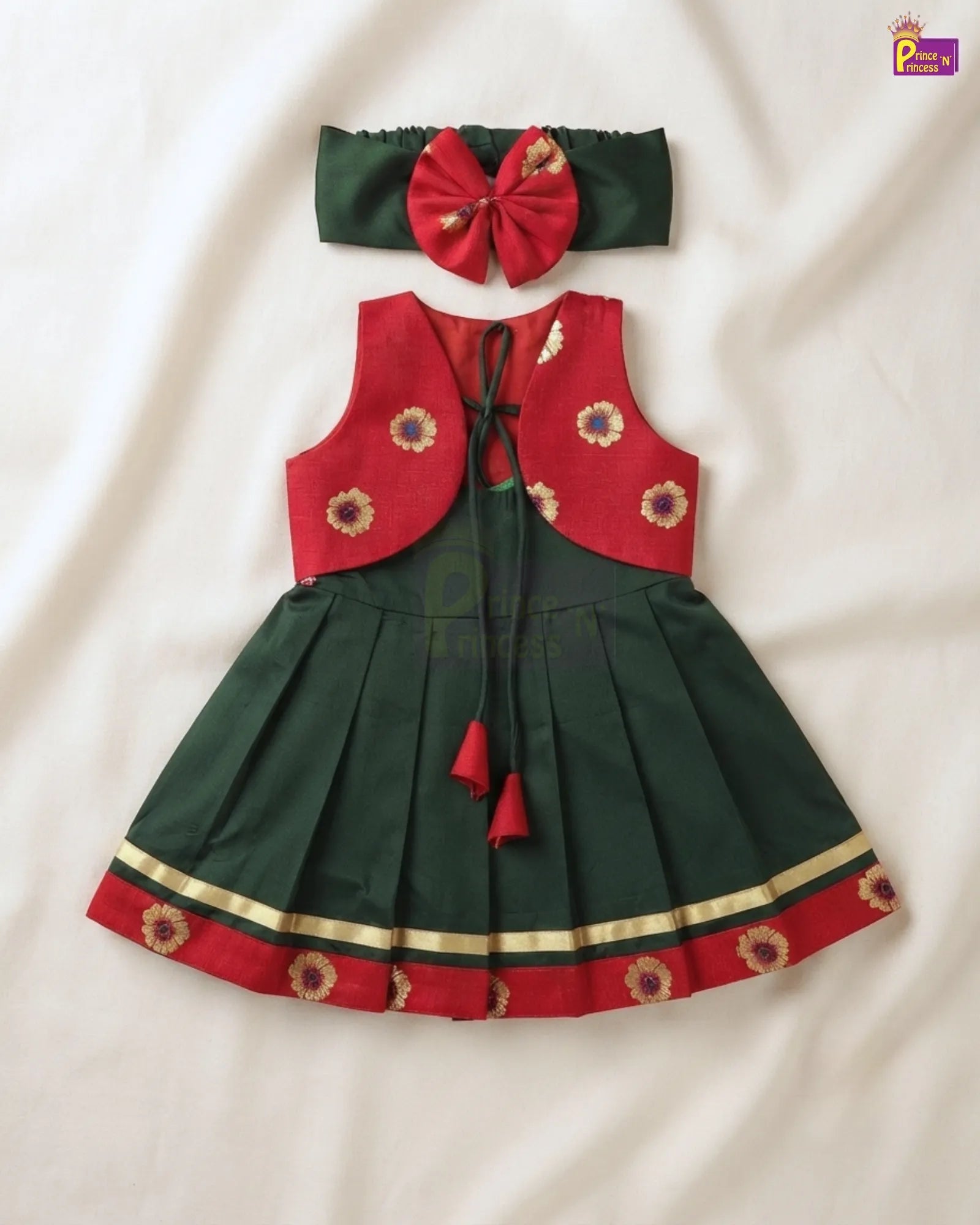 A toddler girl's traditional festive outfit set consisting of a dark green and red dress with embroidered floral details, a matching red bow headband, and a dark green belt, laid flat on a white fabric background.