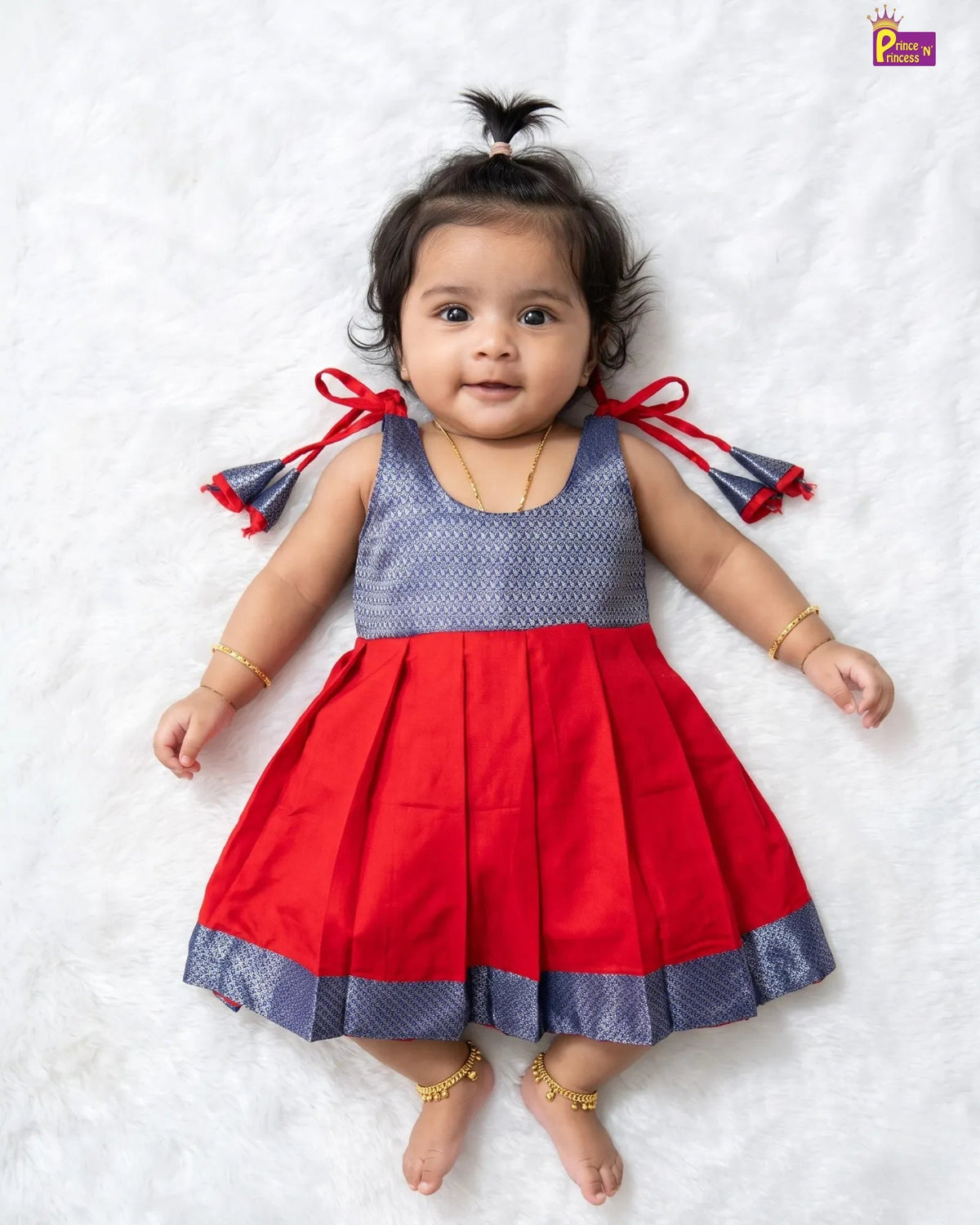 A toddler girl lying on a white textured blanket, wearing a sleeveless red and navy blue dress with a patterned bodice, red bow details on the sides, and a pleated skirt.