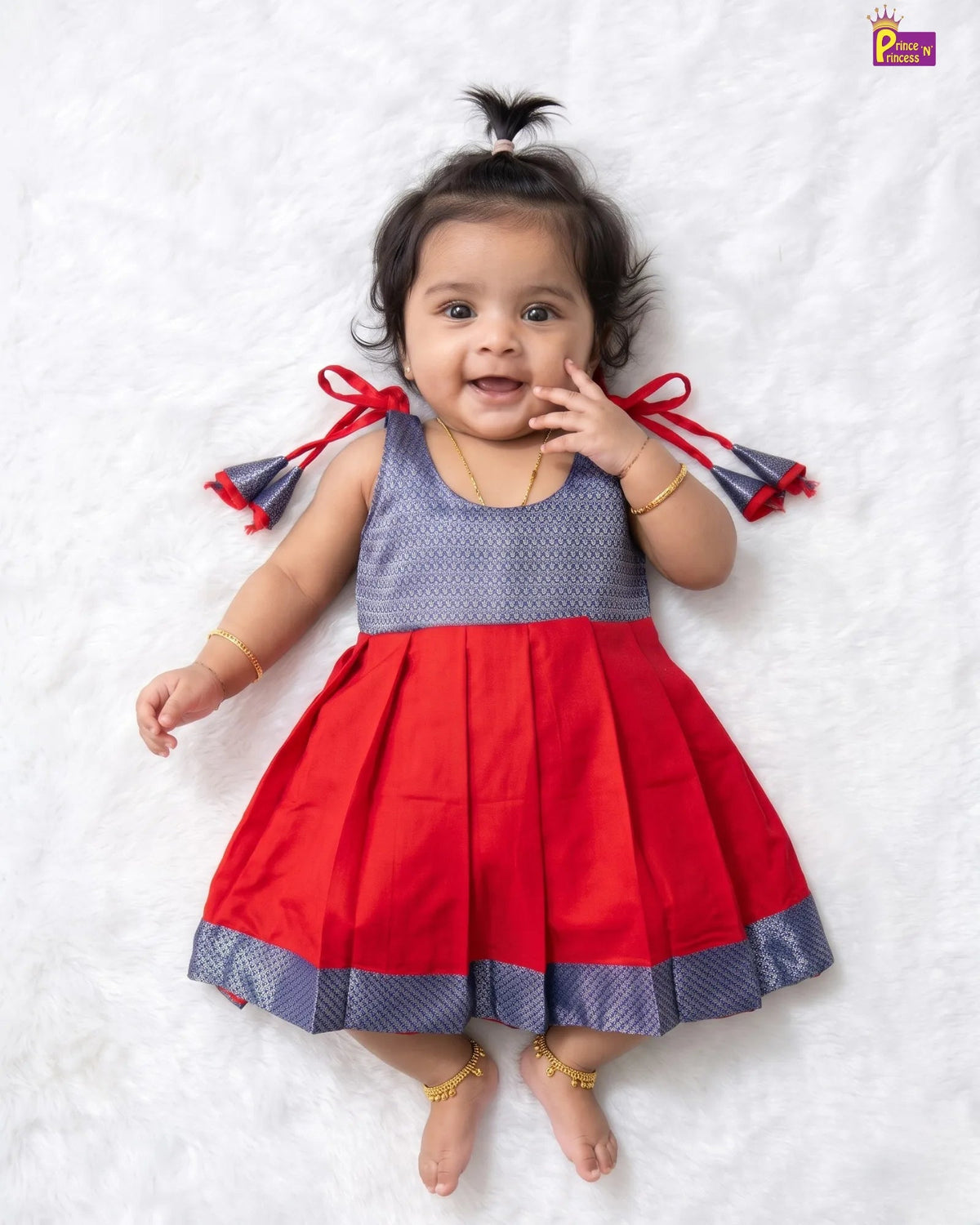 A baby girl lying on a white textured blanket, wearing a sleeveless red and navy blue dress with a patterned bodice, red side bows, and a pleated skirt.