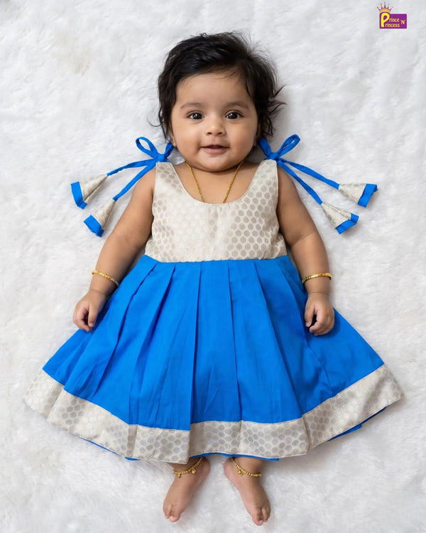 A baby girl wearing a sleeveless blue and cream dress with patterned details, lying on a white textured blanket. The dress features a solid blue pleated skirt with a cream patterned hem and matching blue ribbon bows on the back.