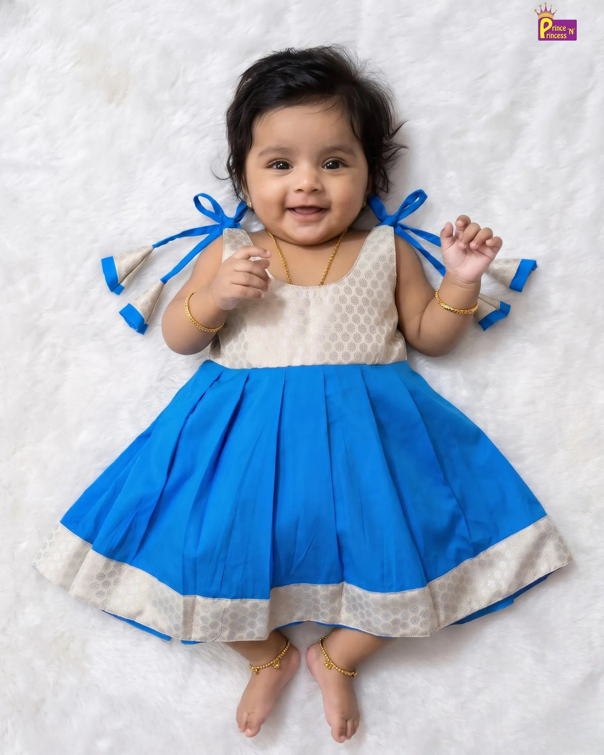A baby girl lying on a white textured blanket, wearing a sleeveless blue and cream dress with blue ribbon bows on the sides. She is wearing gold jewelry, including a necklace, bracelets, and anklets.