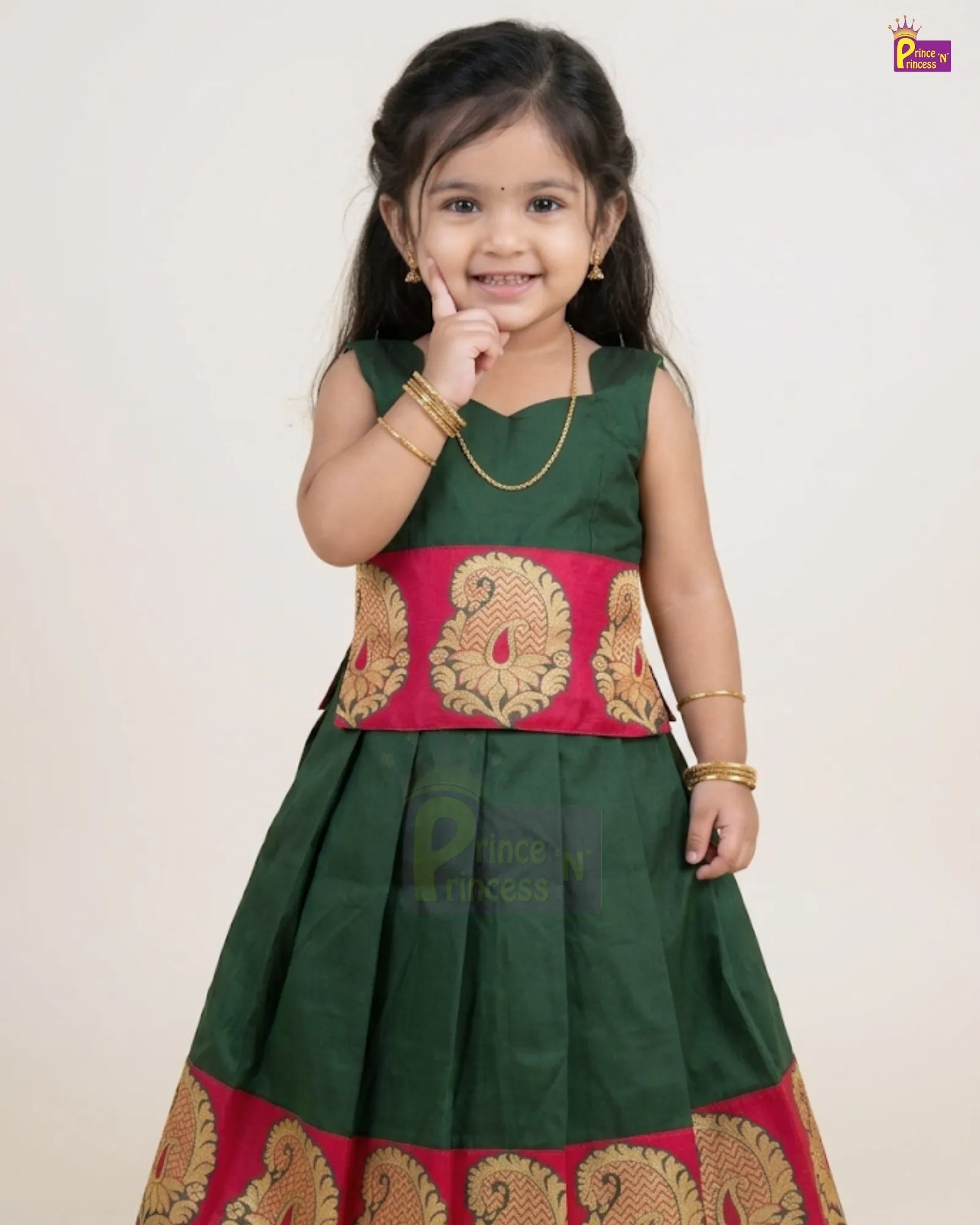 A young girl modeling a traditional Indian dress with a dark green body, a bright pink waistband, and gold paisley patterns, set against a plain light background.