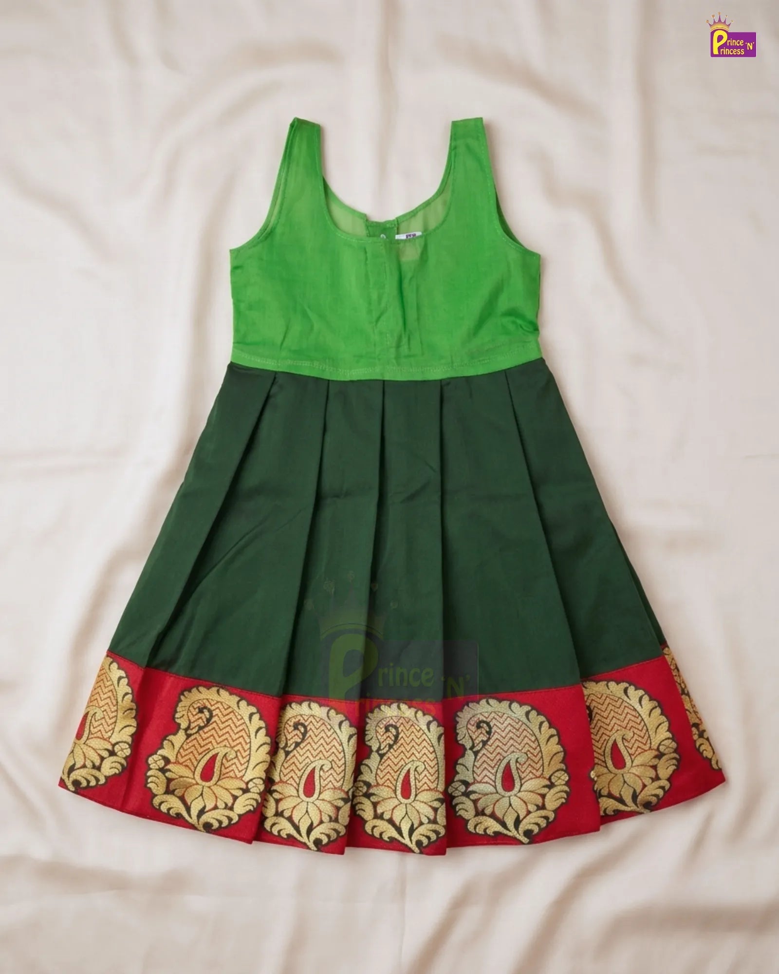 A sleeveless children's dress laid flat on a white fabric background, featuring a bright green bodice, a dark green pleated skirt, and a red border with gold paisley patterns.