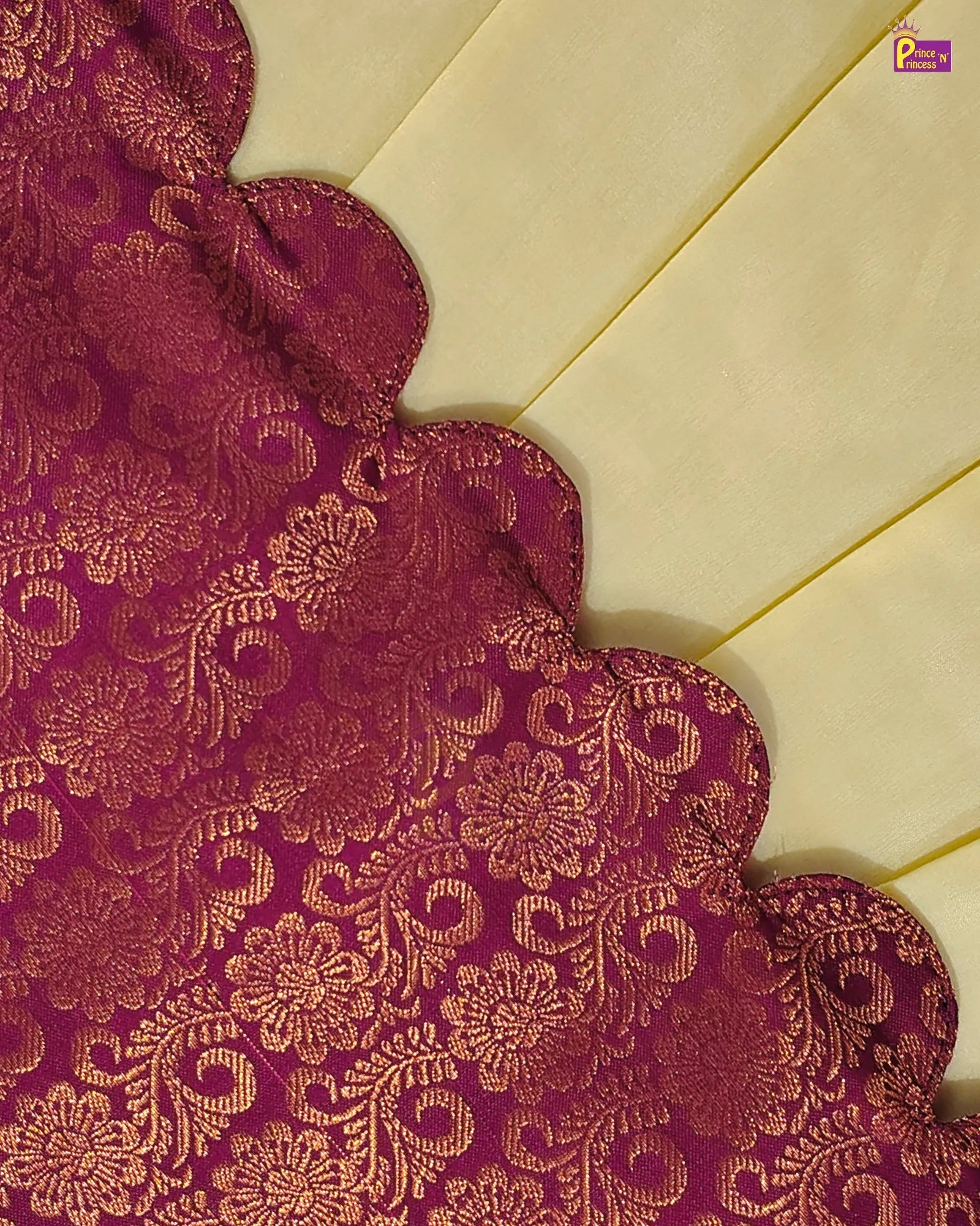 A close-up of a rich magenta-purple brocade fabric with an intricate gold floral and scroll pattern, laid diagonally over a cream-colored background.
