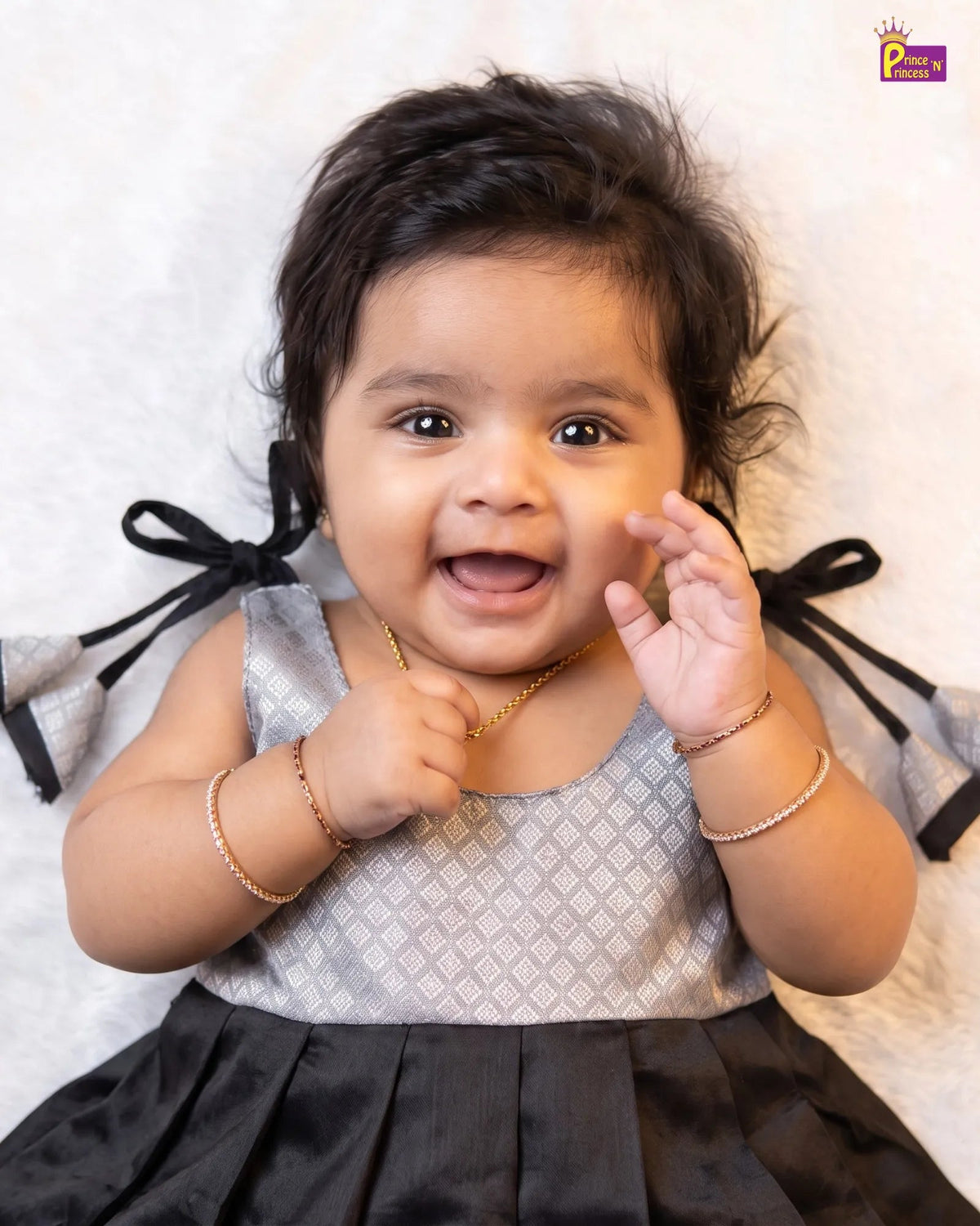 A smiling baby girl wearing a sleeveless silver and black dress with black bow details on the shoulders, lying on a white textured blanket. She is wearing gold bangles.