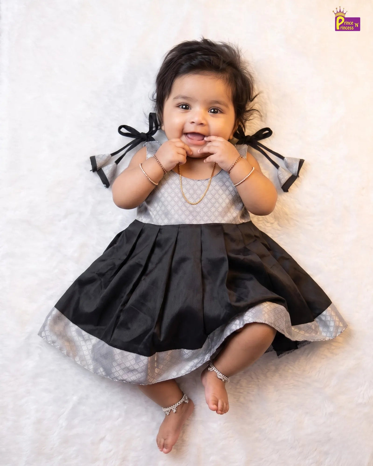 A baby girl wearing a black and silver dress with bow details, lying on a white textured blanket.