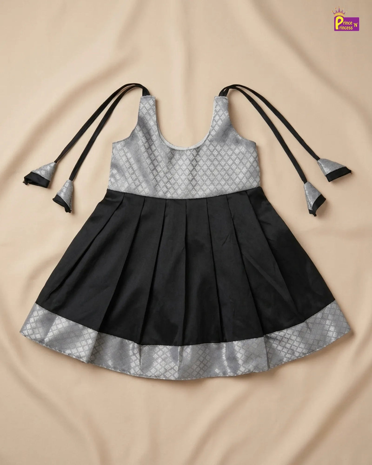 A sleeveless black and silver party dress for girls, featuring a patterned silver top, a pleated black skirt, and long black ties with silver tips, laid flat on a beige fabric background.