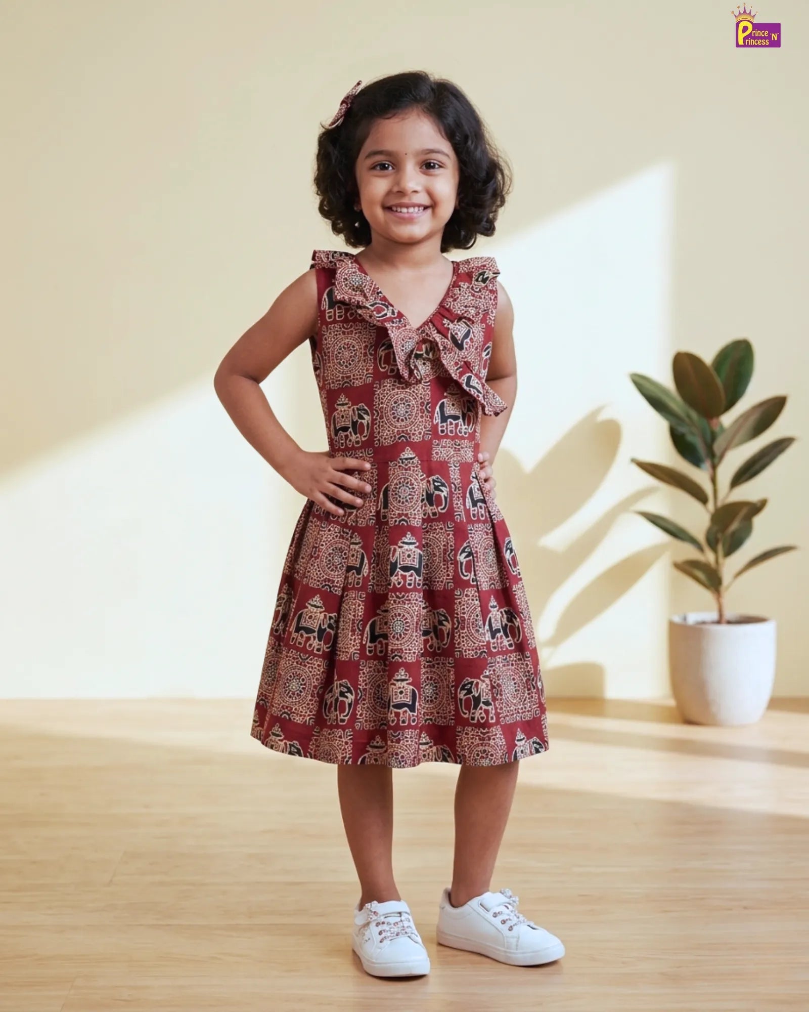 Young girl wearing a patterned dress standing indoors with a plant in the background