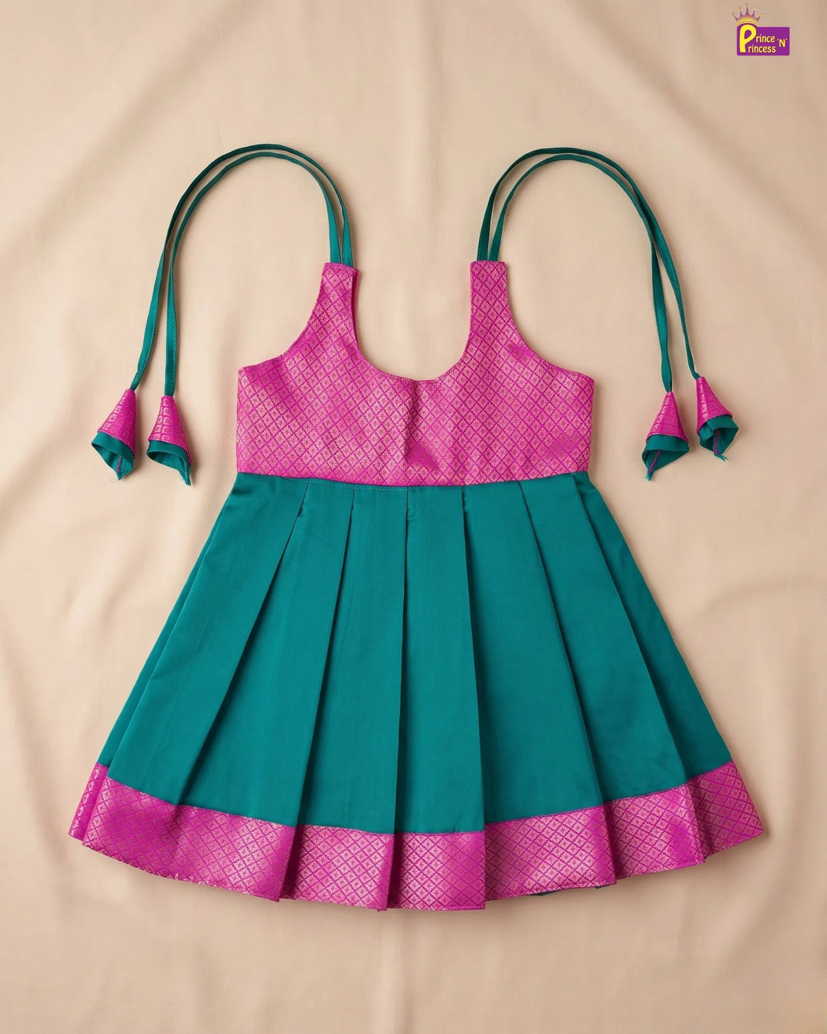 A sleeveless children's dress laid flat on a beige fabric background, featuring a bright pink patterned bodice and hemband with teal-green shoulder ties and a teal-green pleated skirt.