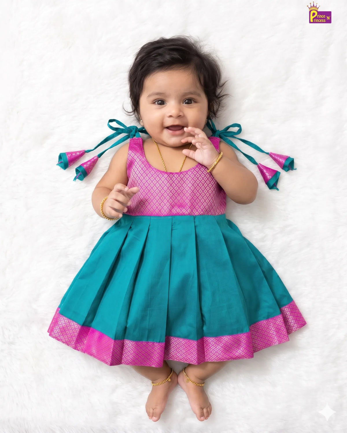 A baby girl lying on a white textured blanket, wearing a sleeveless teal and pink traditional dress with tie-back details on the shoulders and a pleated skirt.
