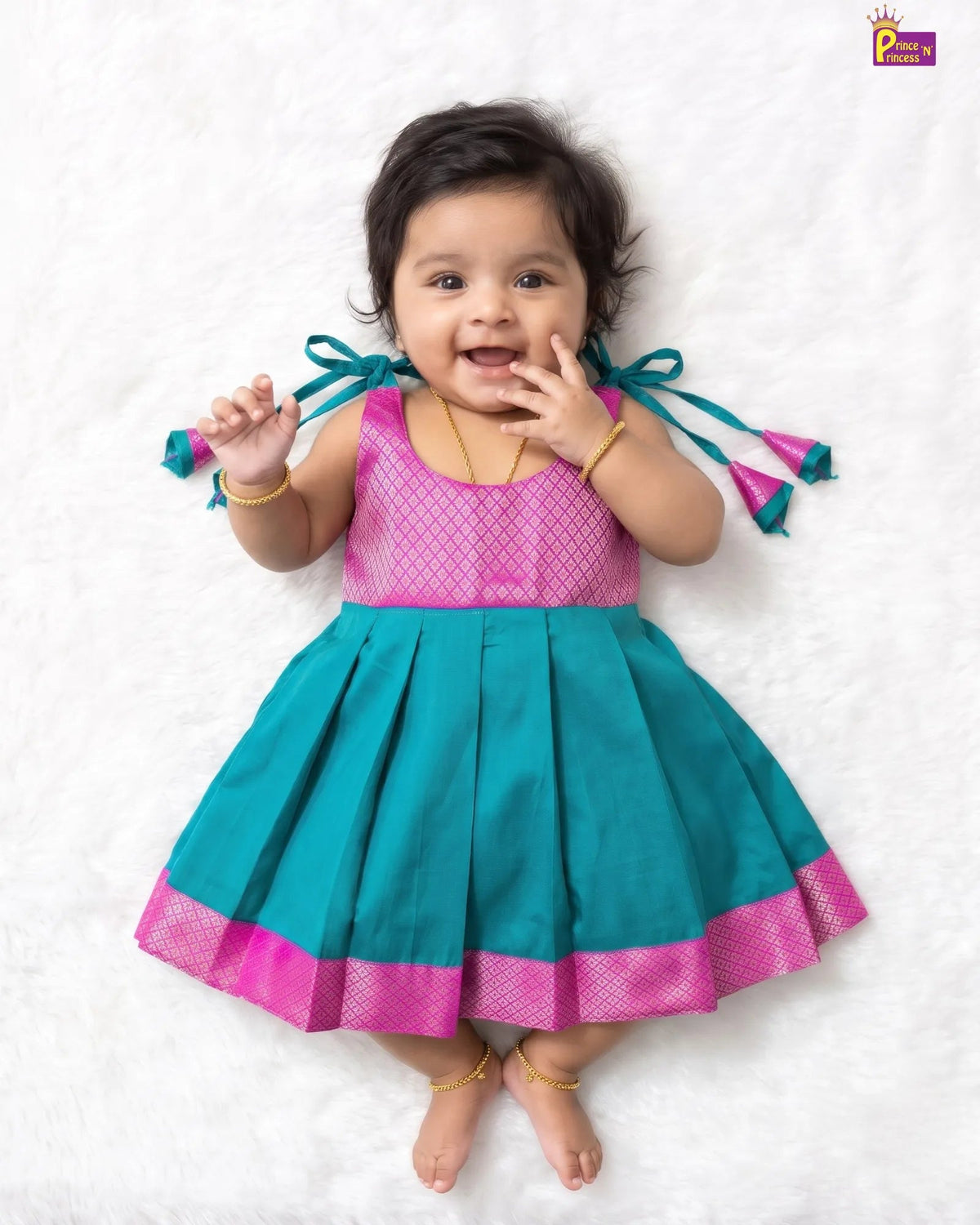 A baby girl wearing a traditional Indian dress with a pink mesh bodice and a teal pleated skirt with a pink border, lying on a white textured blanket.
