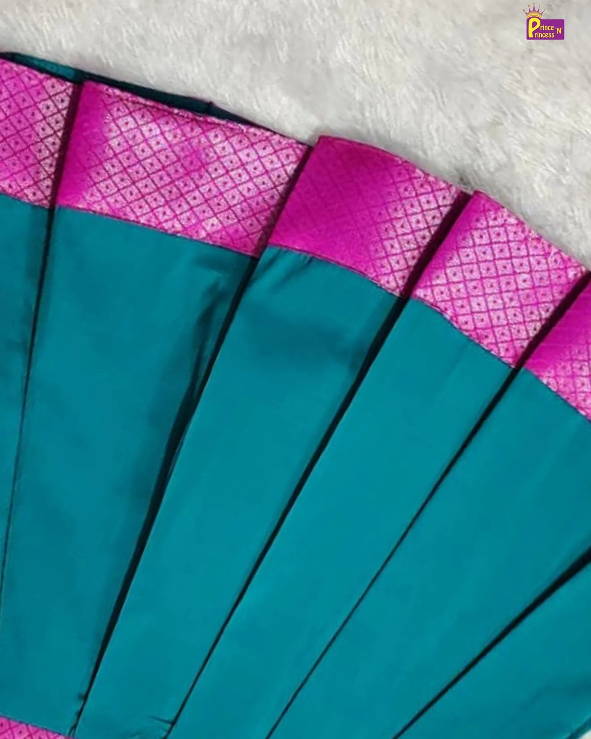 A close-up of a teal and magenta fabric accessory, likely a sari or scarf, laid flat on a white textured surface. The teal body is solid, while the magenta border features a woven geometric pattern.
