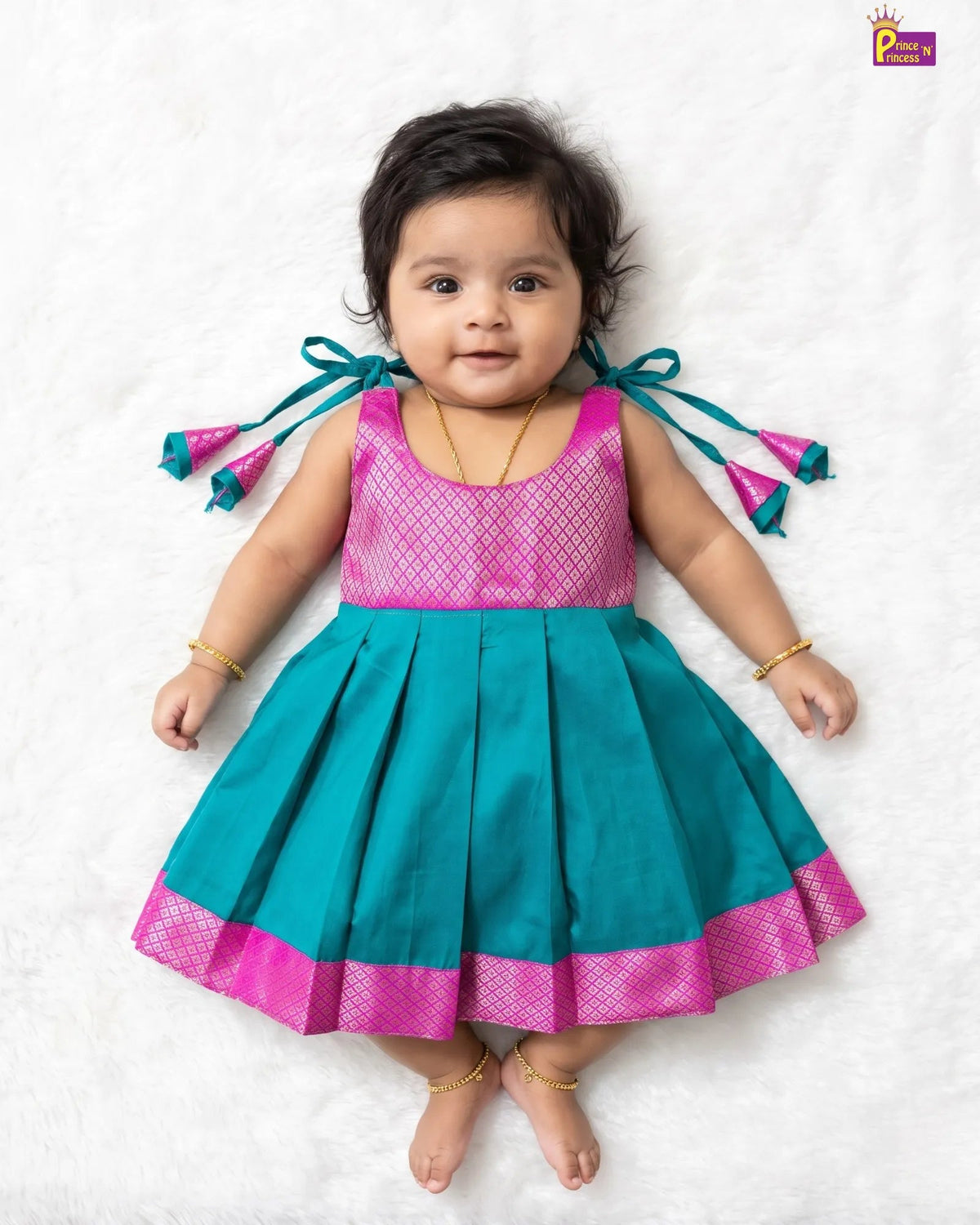 A baby girl wearing a sleeveless teal and pink traditional dress with pleated skirt and bow details, lying on a white textured blanket.