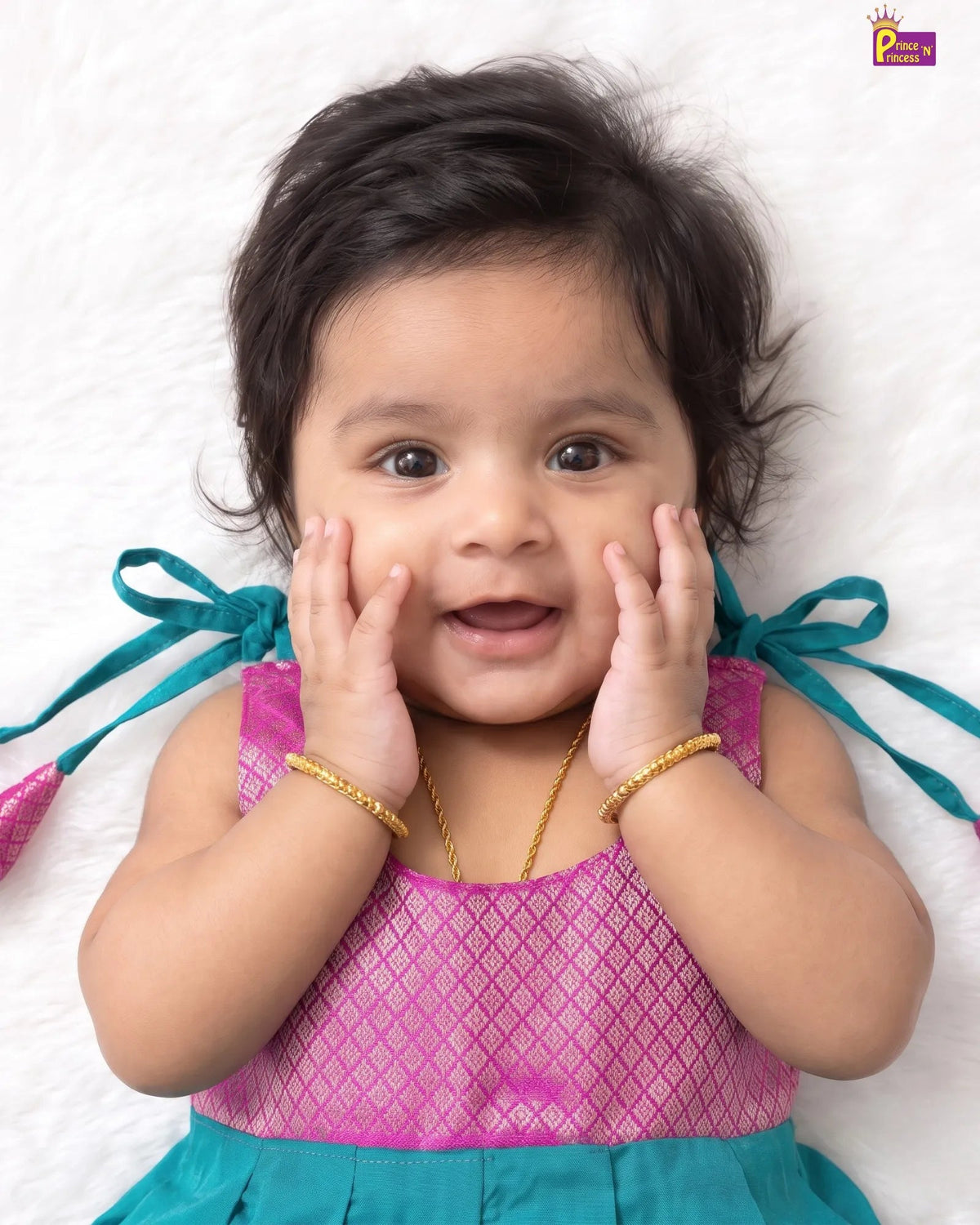 A baby girl lying on a white textured blanket, wearing a pink and teal dress with gold jewelry.