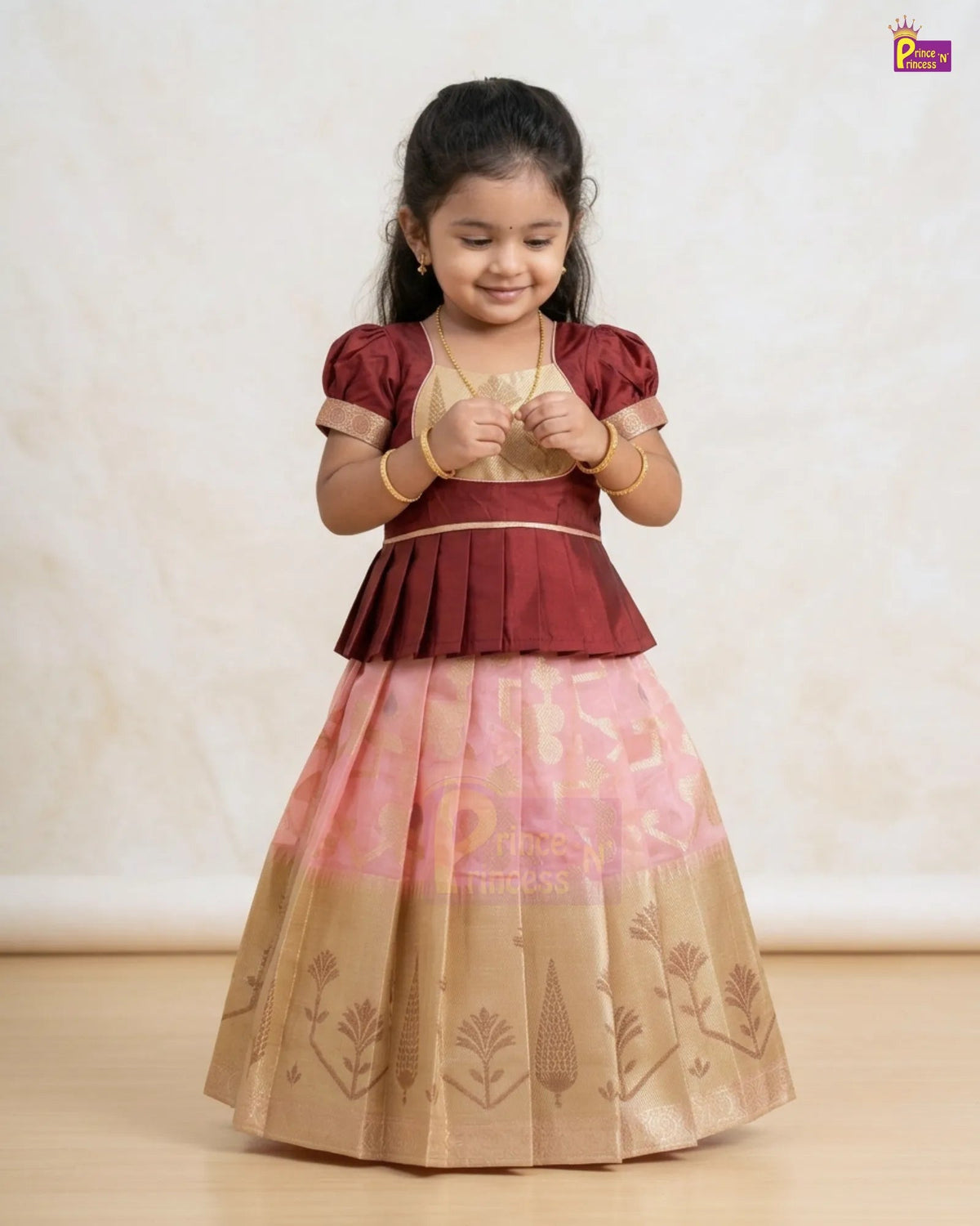 A young girl modeling a traditional Indian ethnic dress with a maroon peplum-style bodice, a light pink upper skirt, and a beige lower skirt with printed motifs, set against a neutral cream background.