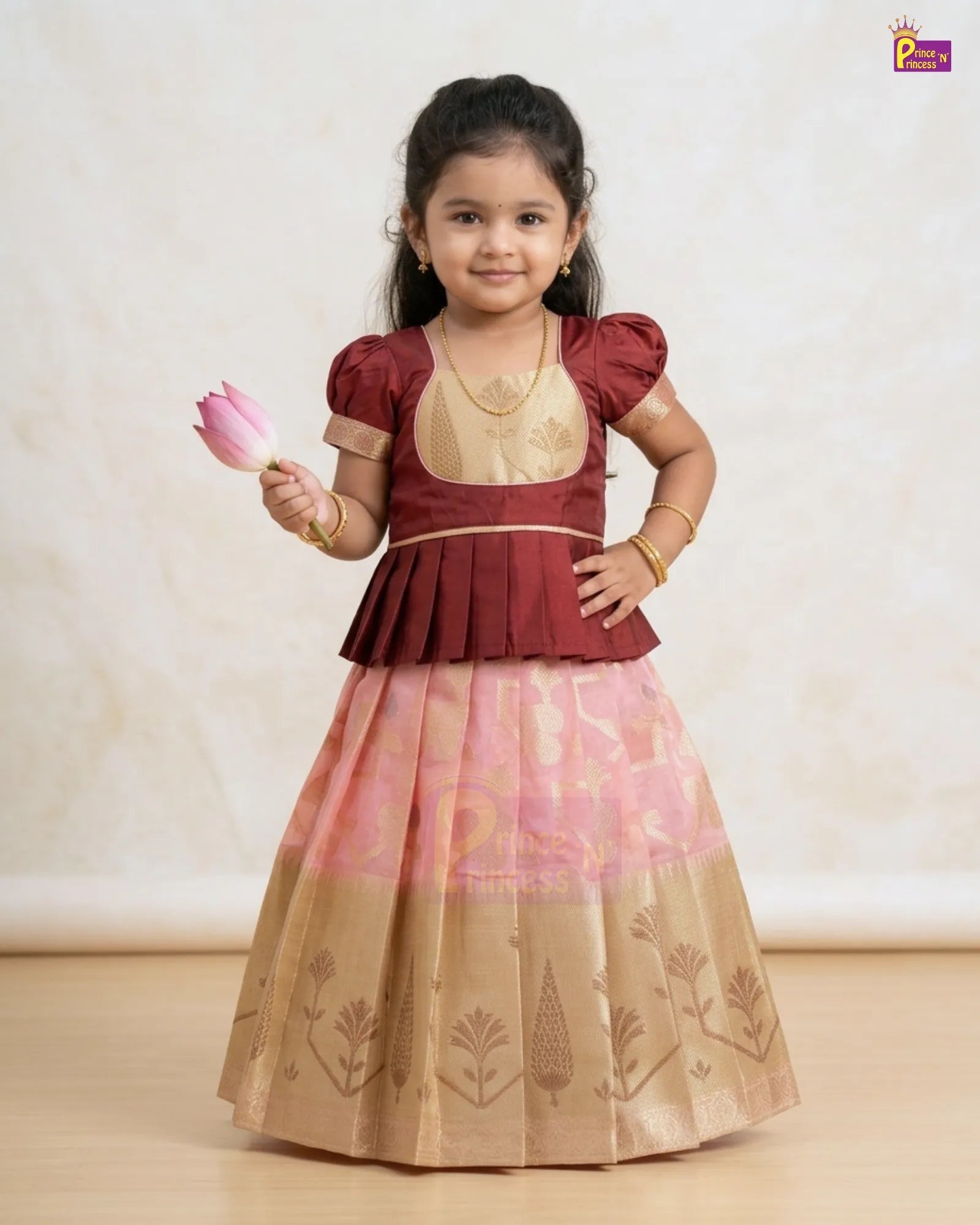 A young girl modeling a traditional Indian outfit consisting of a maroon and gold blouse with a pink and gold patterned skirt, holding a pink lotus flower against a neutral beige background.