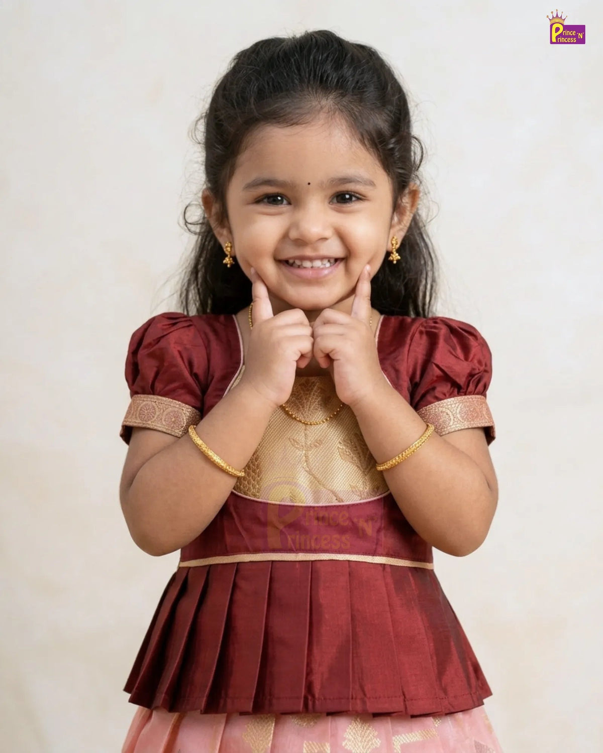 A young girl smiling and posing in a traditional Indian outfit. The outfit features a maroon top with gold embroidery and a pleated skirt in pink and gold.