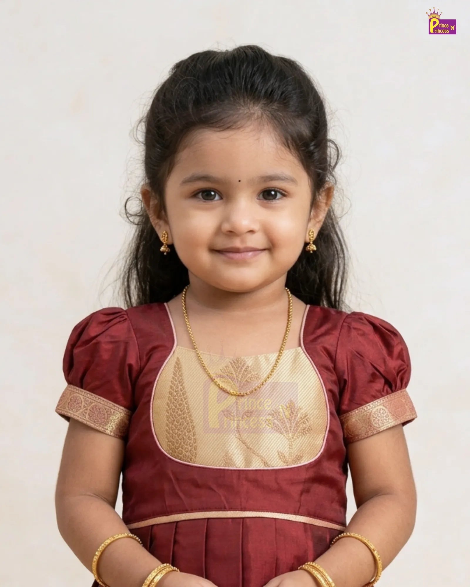 A young girl with dark hair wearing a traditional Indian maroon and gold dress, styled with gold jewelry, photographed against a plain light background.