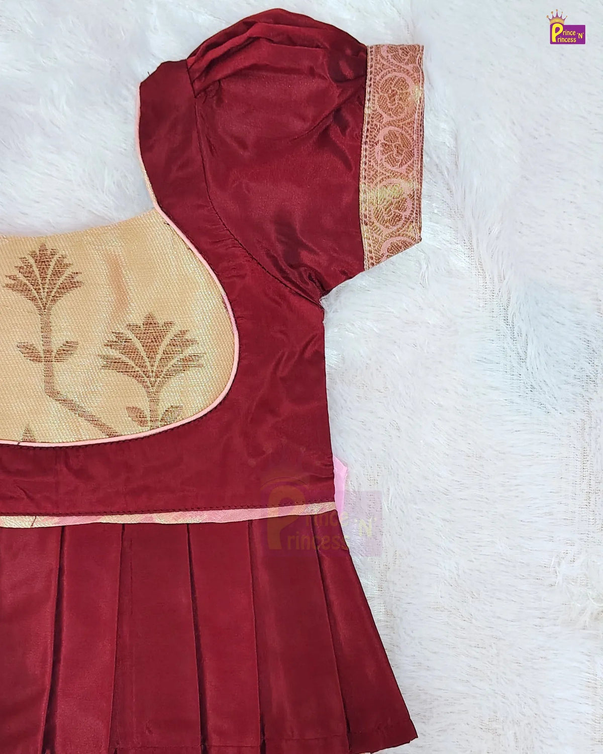 A close-up of a traditional Indian outfit, likely a salwar suit or kurta set, featuring a deep maroon red fabric with a pleated skirt, a beige embroidered yoke with gold motifs, and a patterned gold and red border on the sleeve, laid flat on a white textured background.