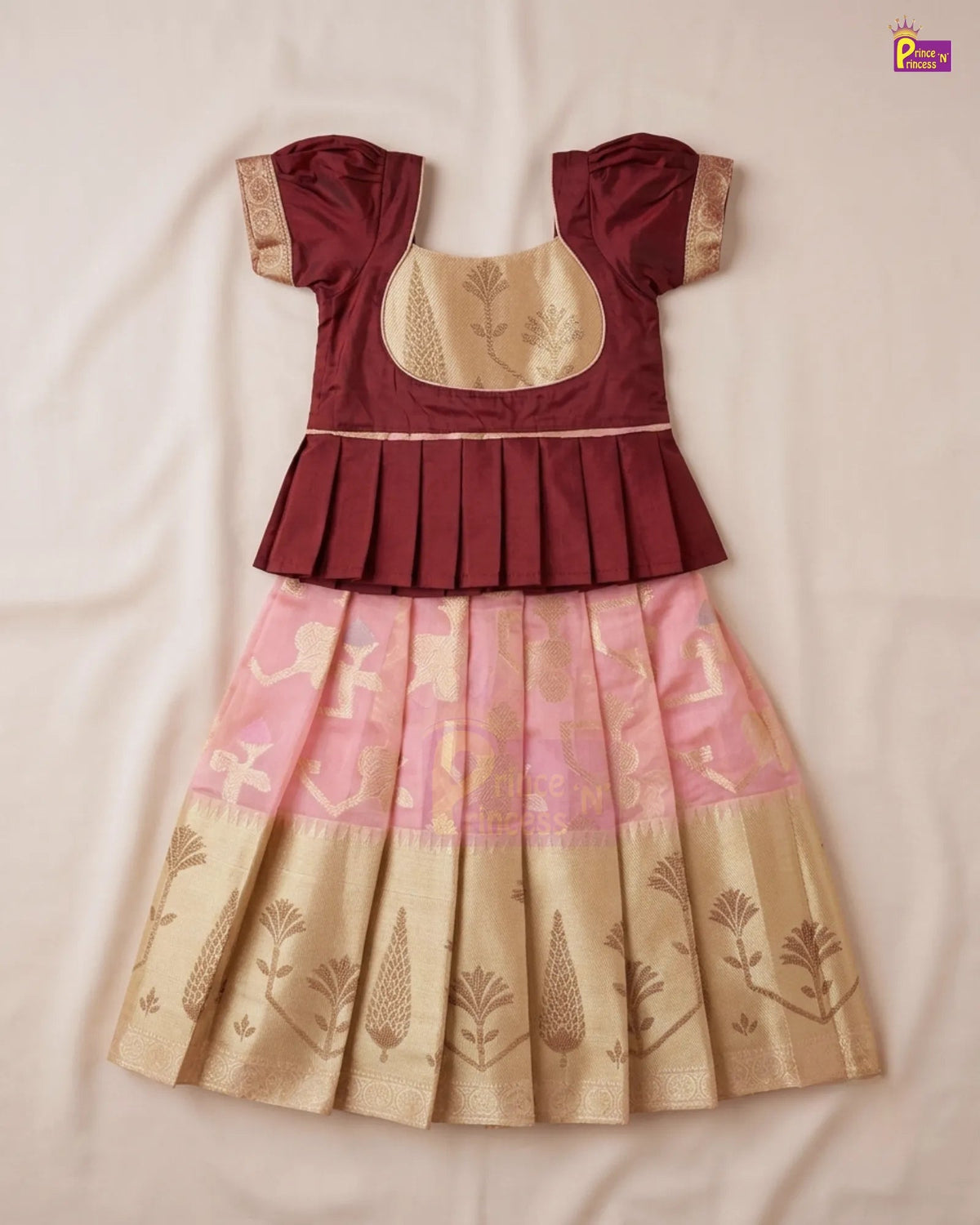 A traditional Indian girl's dress with a maroon peplum top, beige embroidered bodice, and a pleated skirt in pink and beige with gold zari motifs, laid flat on a light beige fabric background.