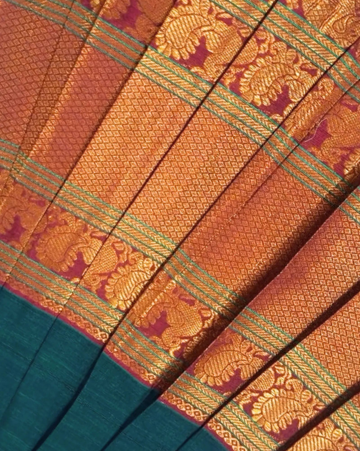 Close-up of a folded textile with a richly patterned golden-orange body, purple border details, and a teal lower section with woven ornamental stripes.