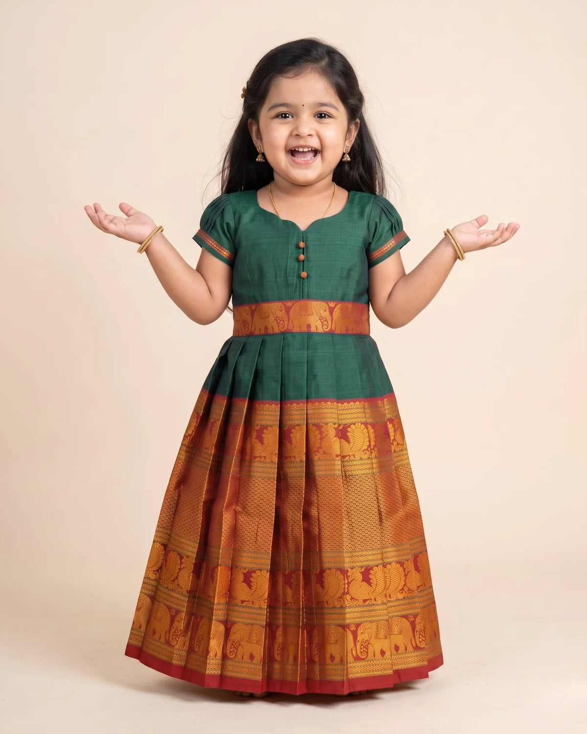 A young girl smiling with her hands outstretched, wearing a traditional Indian dress with a dark green bodice and a pleated skirt featuring gold and orange patterns.