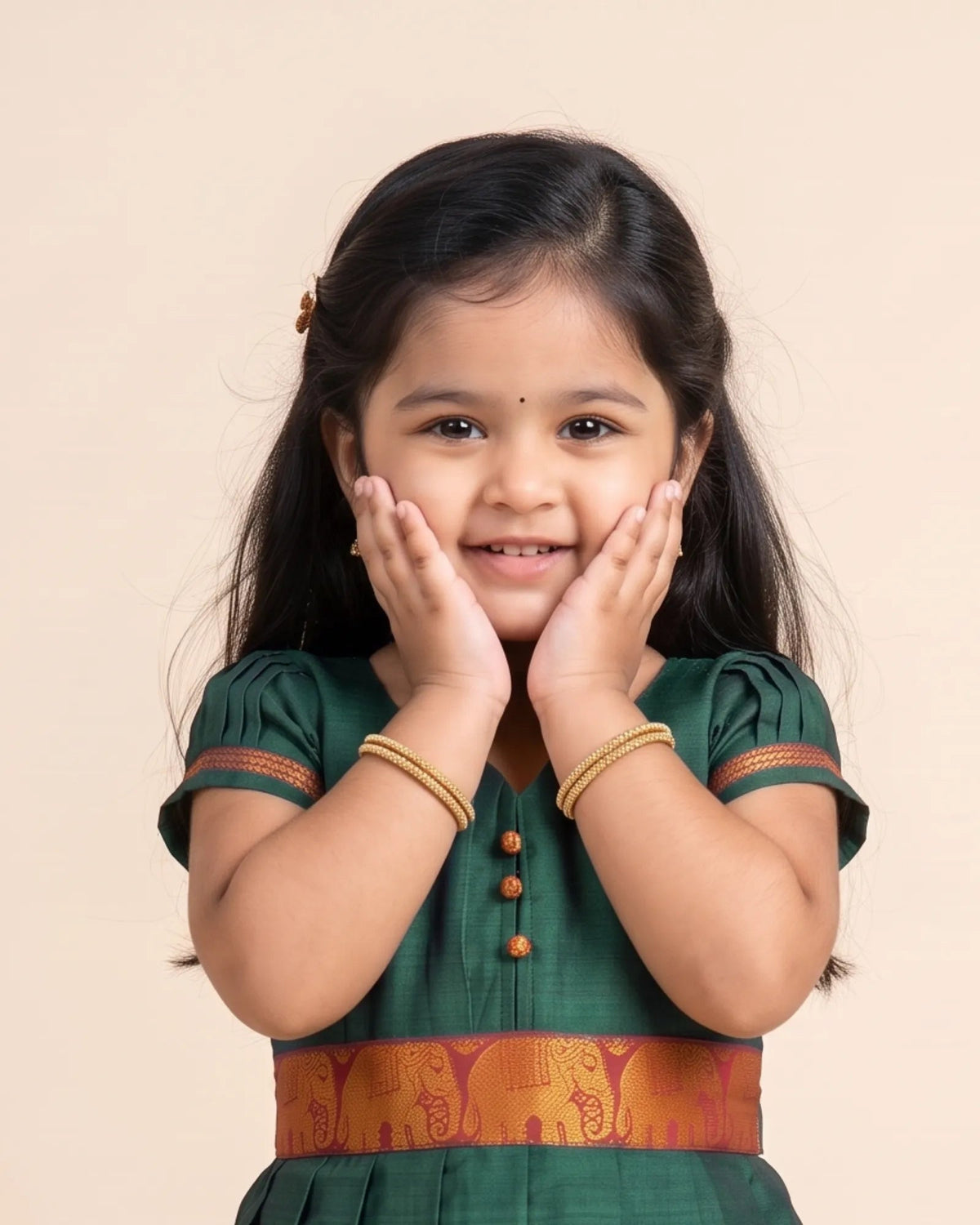 A young girl with dark hair wearing a dark green traditional outfit with gold accents and bangles, posing with her hands on her cheeks against a solid beige background.