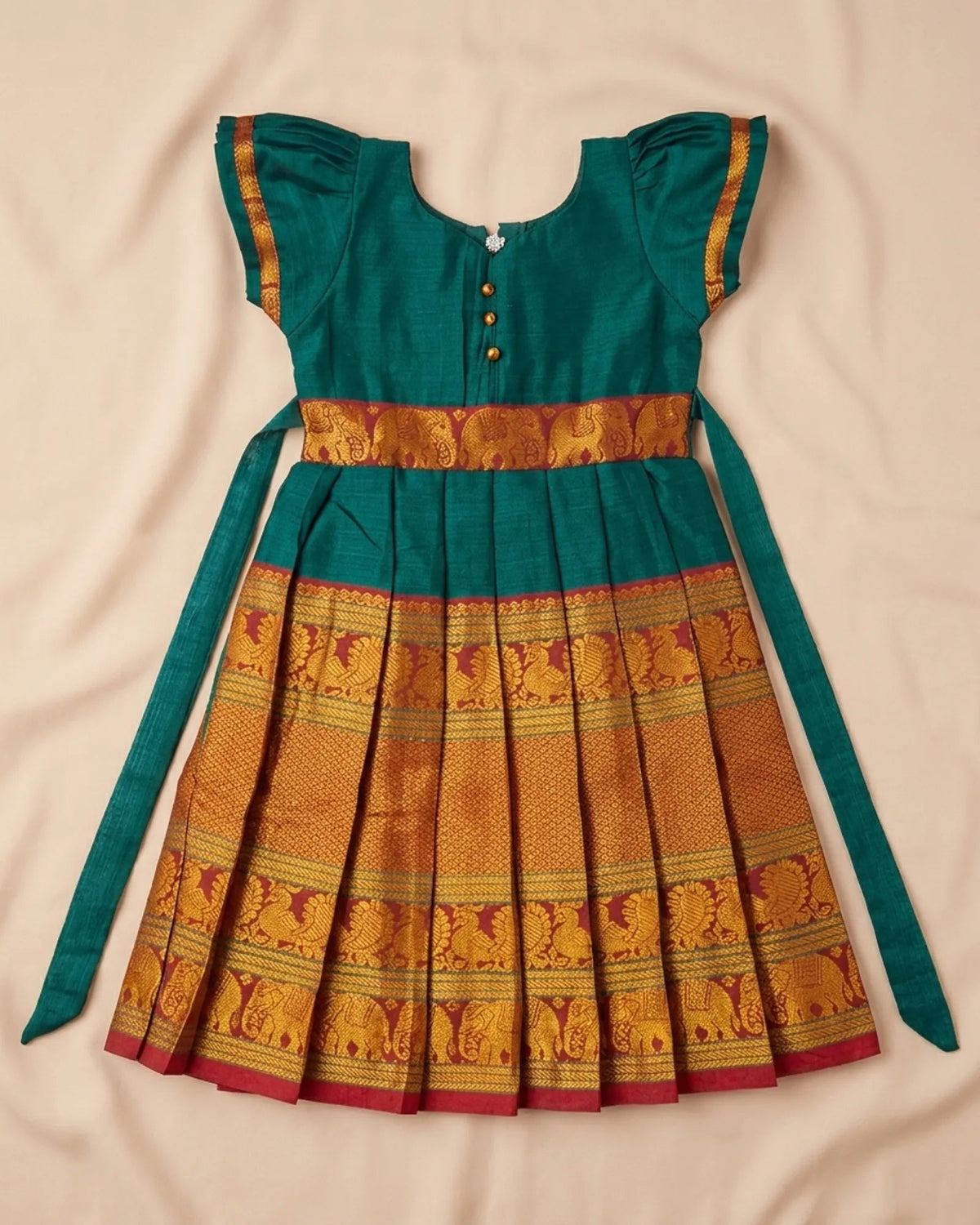 A traditional Indian girl's dress in teal green and gold Banarasi silk, featuring a pleated skirt with an elephant border pattern, a gold waistband, and matching green tie straps, laid flat on a beige fabric background.