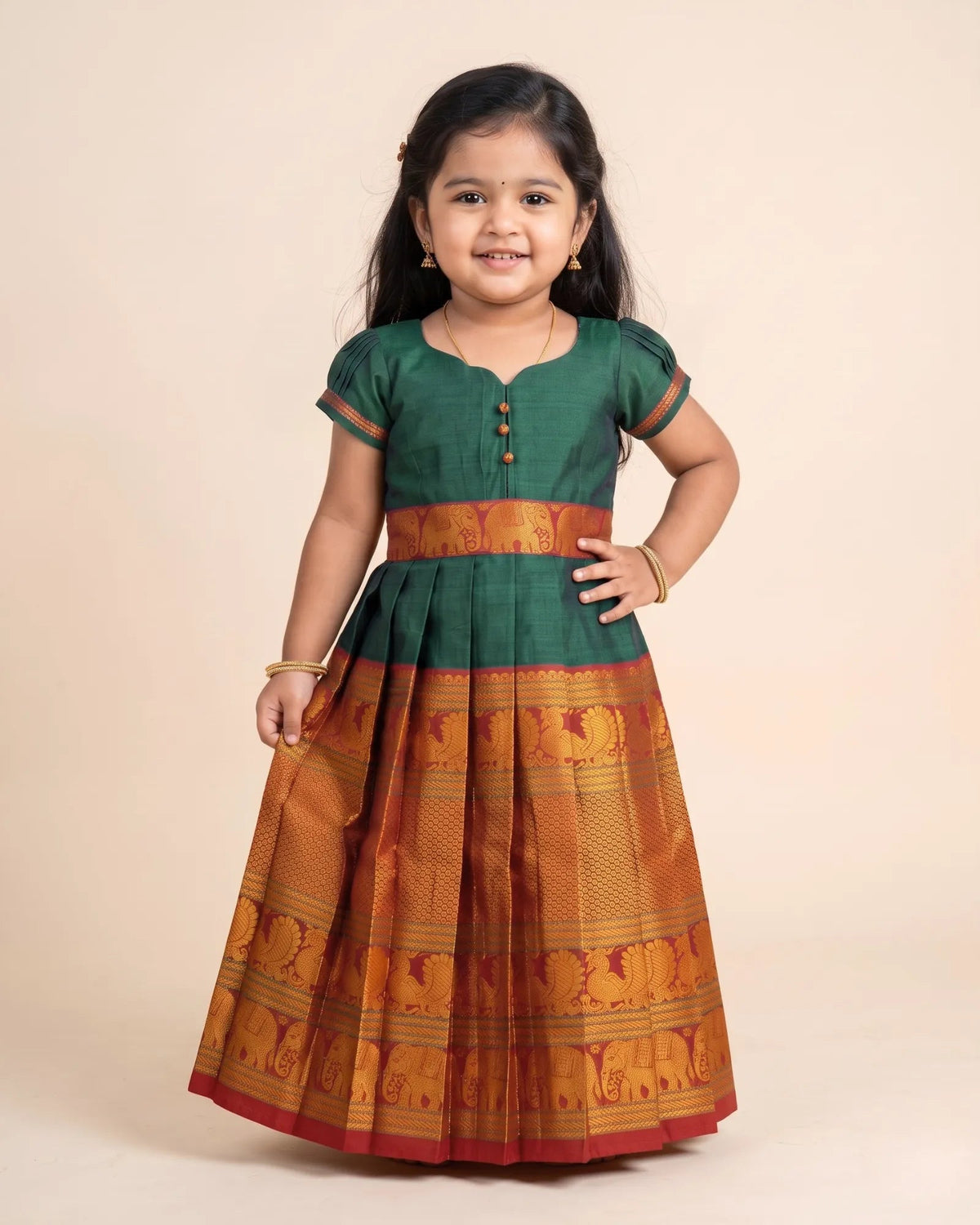 A young girl modeling a traditional Indian dress with a dark green blouse and a pleated skirt featuring a wide orange and gold patterned border, set against a solid beige background.