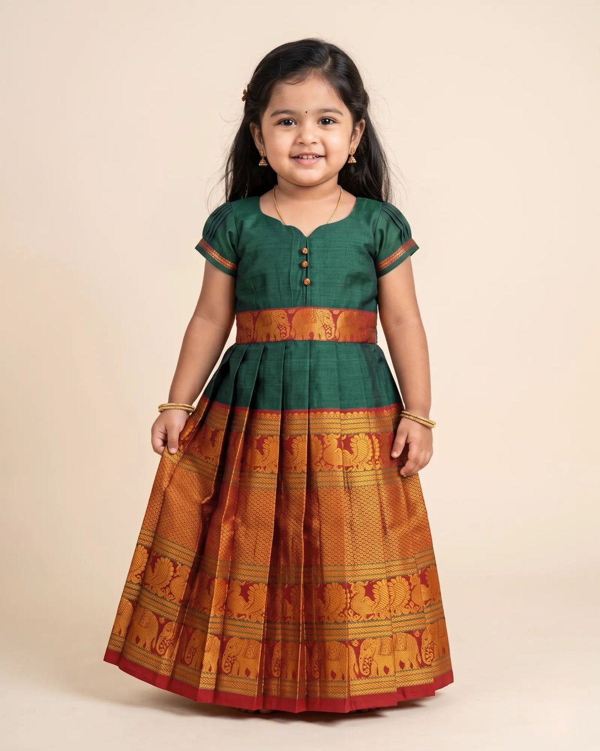 A young girl modeling a traditional Indian dress with a dark green blouse and a pleated orange-gold skirt featuring elephant motifs, set against a solid beige background.