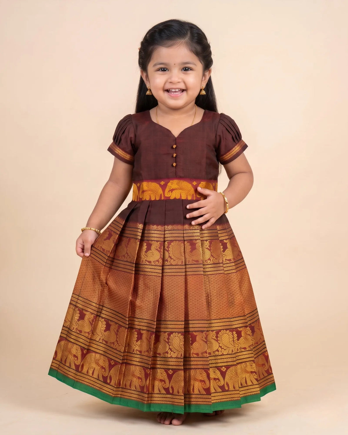 A young girl modeling a traditional Indian dress with a dark brown bodice and a flared golden-brown skirt featuring elephant motifs and a green border, set against a solid beige background.