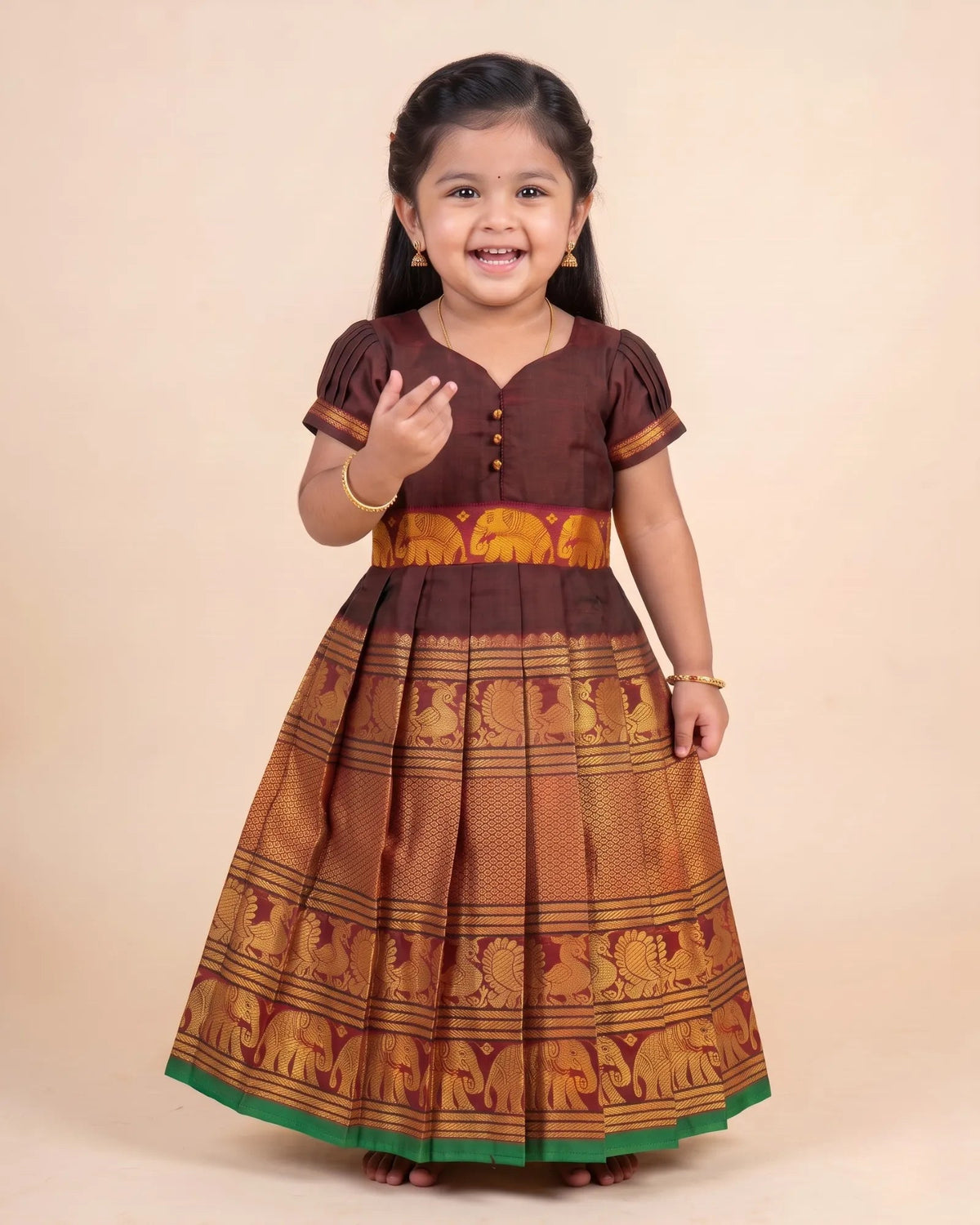 A young girl smiling and posing in a traditional Indian dress. The dress features a dark brown short-sleeve bodice with button details, a wide orange and gold patterned waistband, and a pleated skirt with gold elephant motifs and a green border.