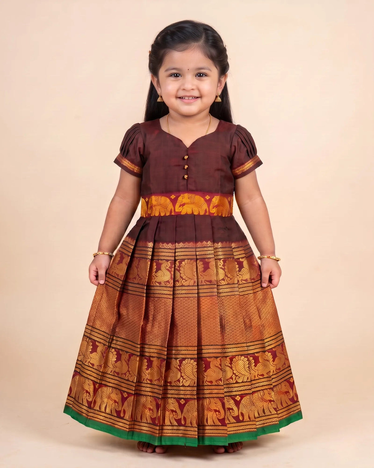 A young girl modeling a traditional Indian dress with a dark brown bodice, gold and orange patterned skirt, and a green hem, set against a plain beige background.