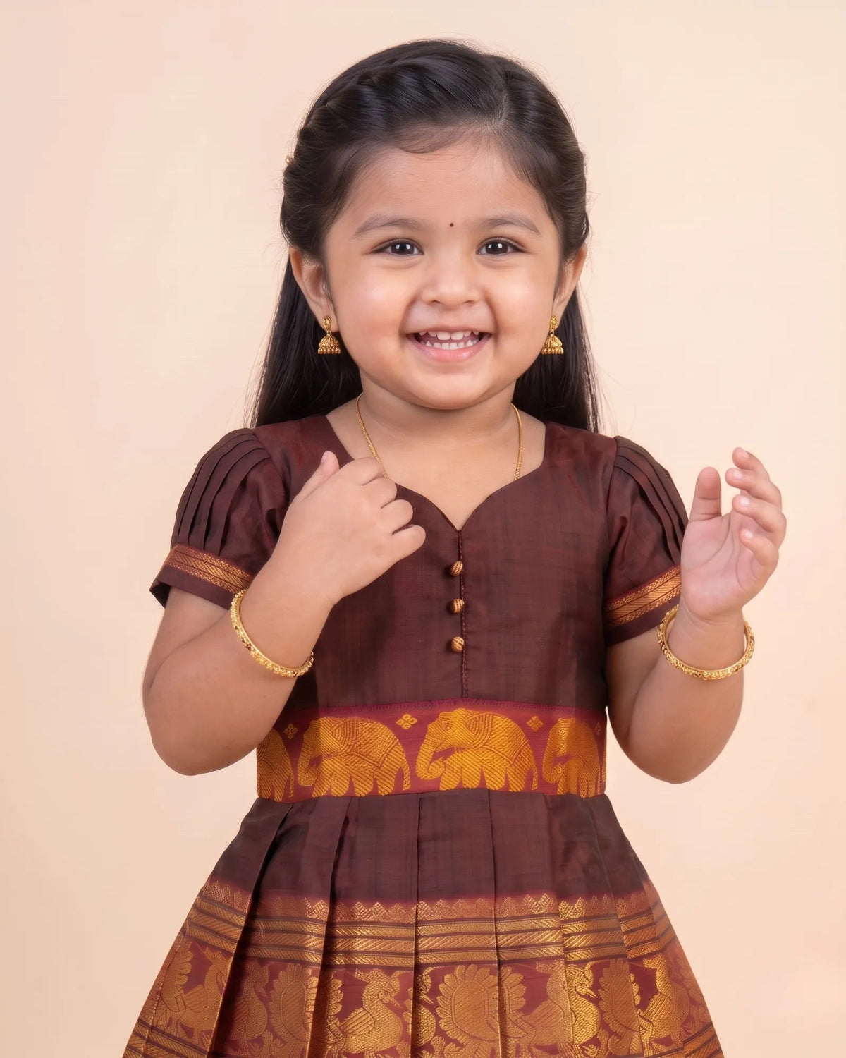 A young girl smiling and wearing a traditional brown and gold Indian dress with gold jewelry, set against a plain beige background.