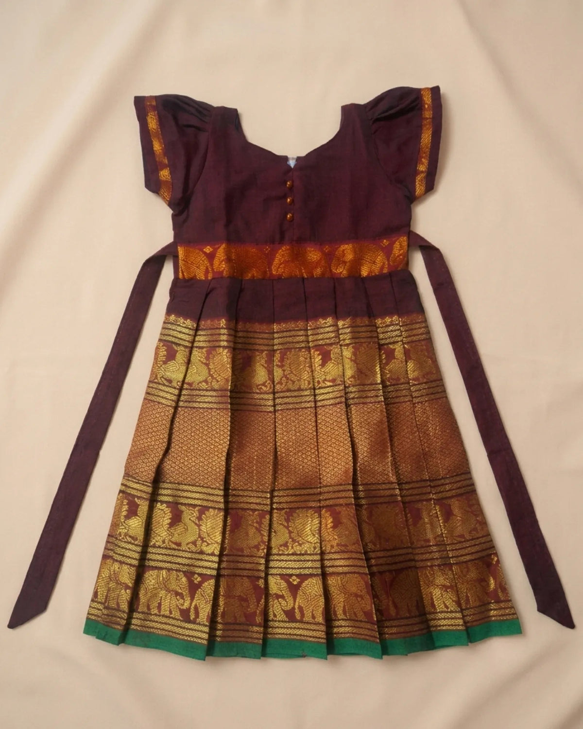A traditional Indian girl's dress laid flat on a beige fabric background. The dress features a deep purple bodice with gold trim, a gold and brown patterned waistband, and a pleated skirt with matching purple and gold patterns and a green border.