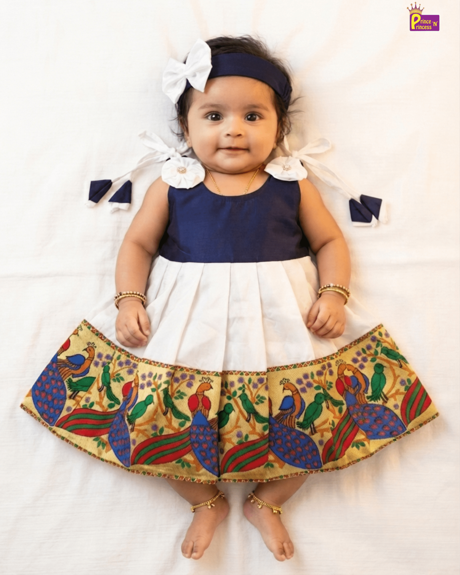 A baby girl lying flat on a white sheet wearing a navy blue and white two-tone dress with a colorful peacock patterned skirt, a navy headband with a white bow, and gold bangles and ankle bracelets.